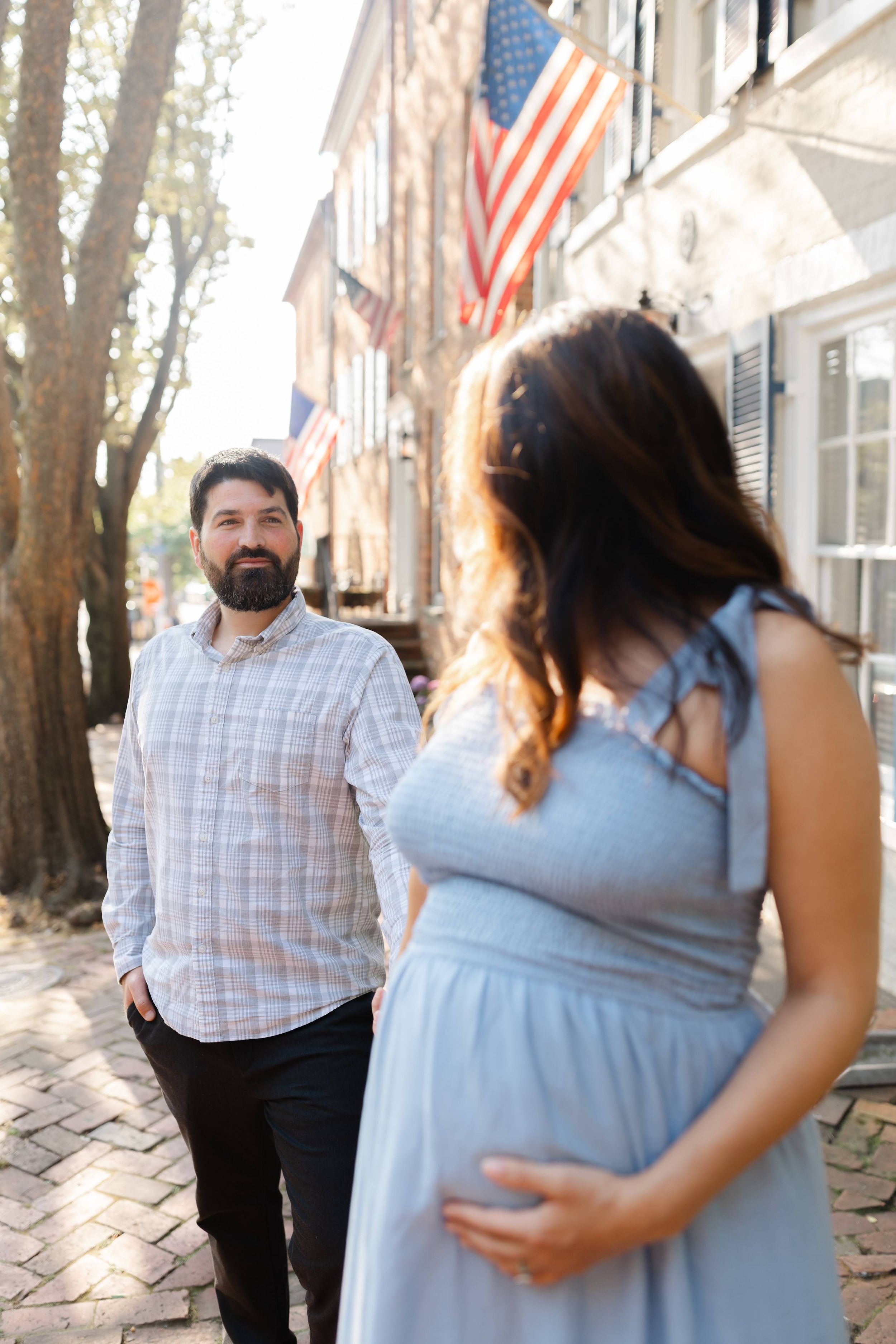 A bearded man smiles at a pregnant woman in a blue dress on a brick sidewalk with American flags in the background.