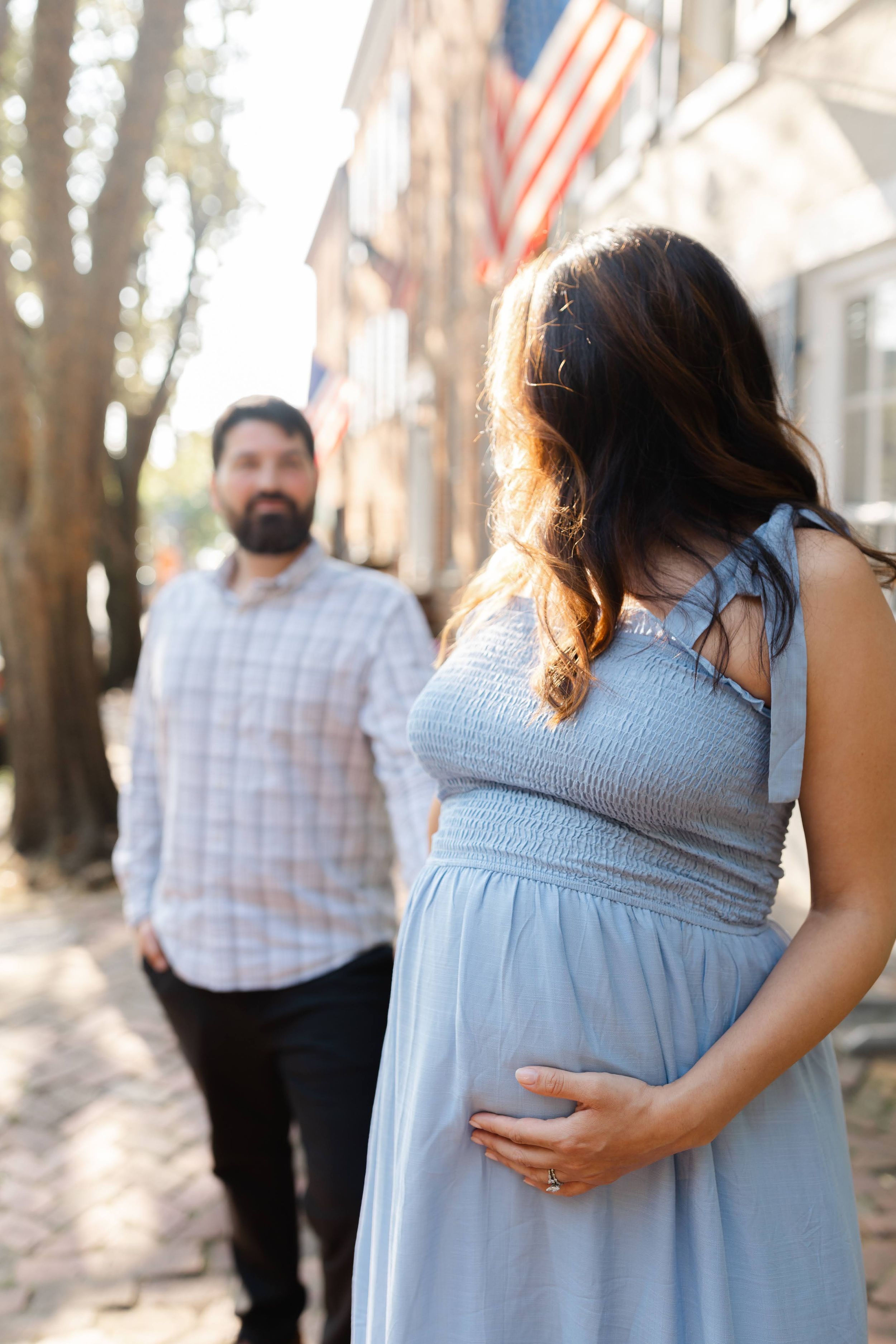 Pregnant woman in blue dress holds her belly outdoors while bearded man stands behind her near brick building with American flag.