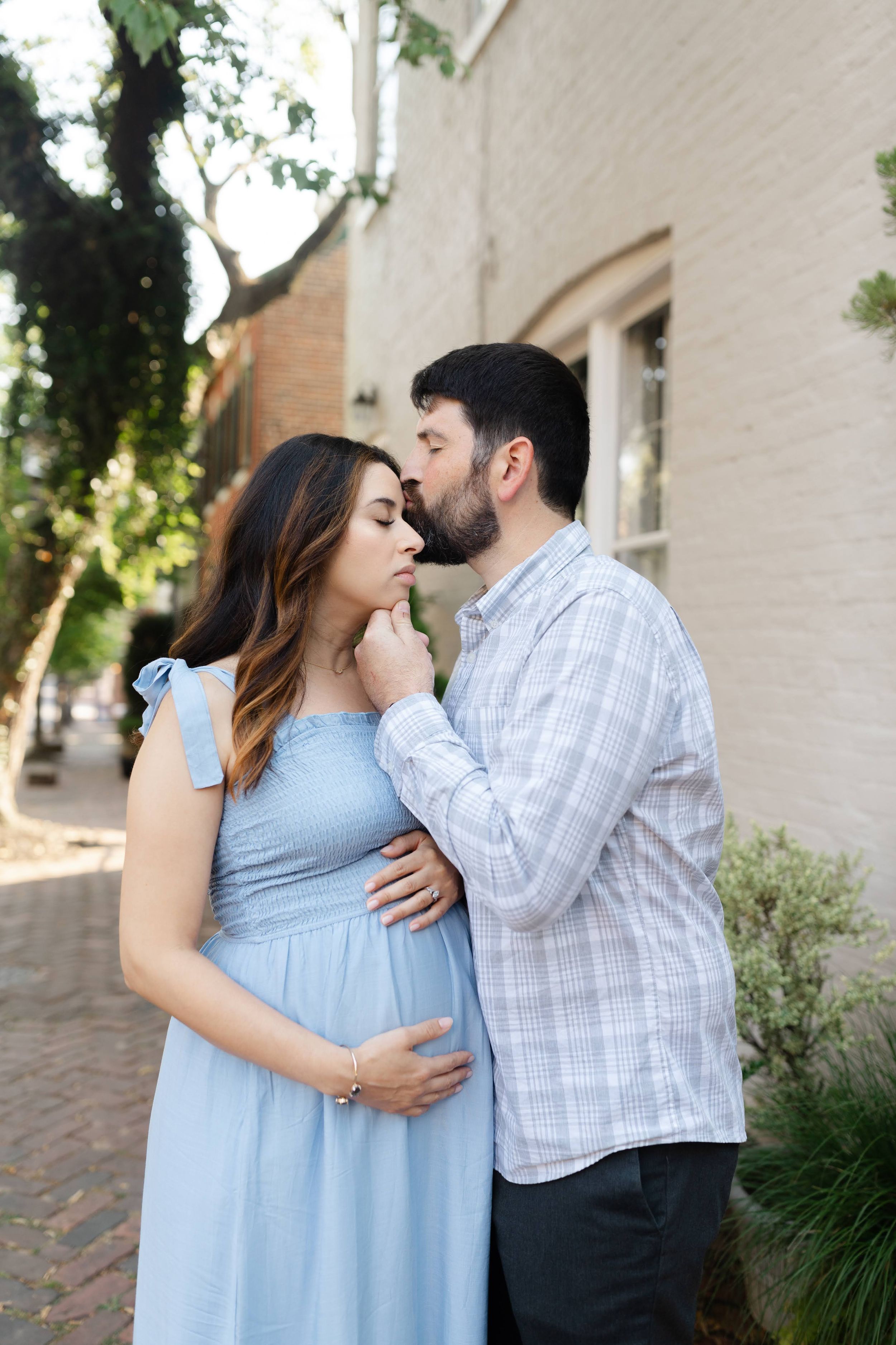 Pregnant woman in blue dress with partner kissing her cheek outdoors near brick building.