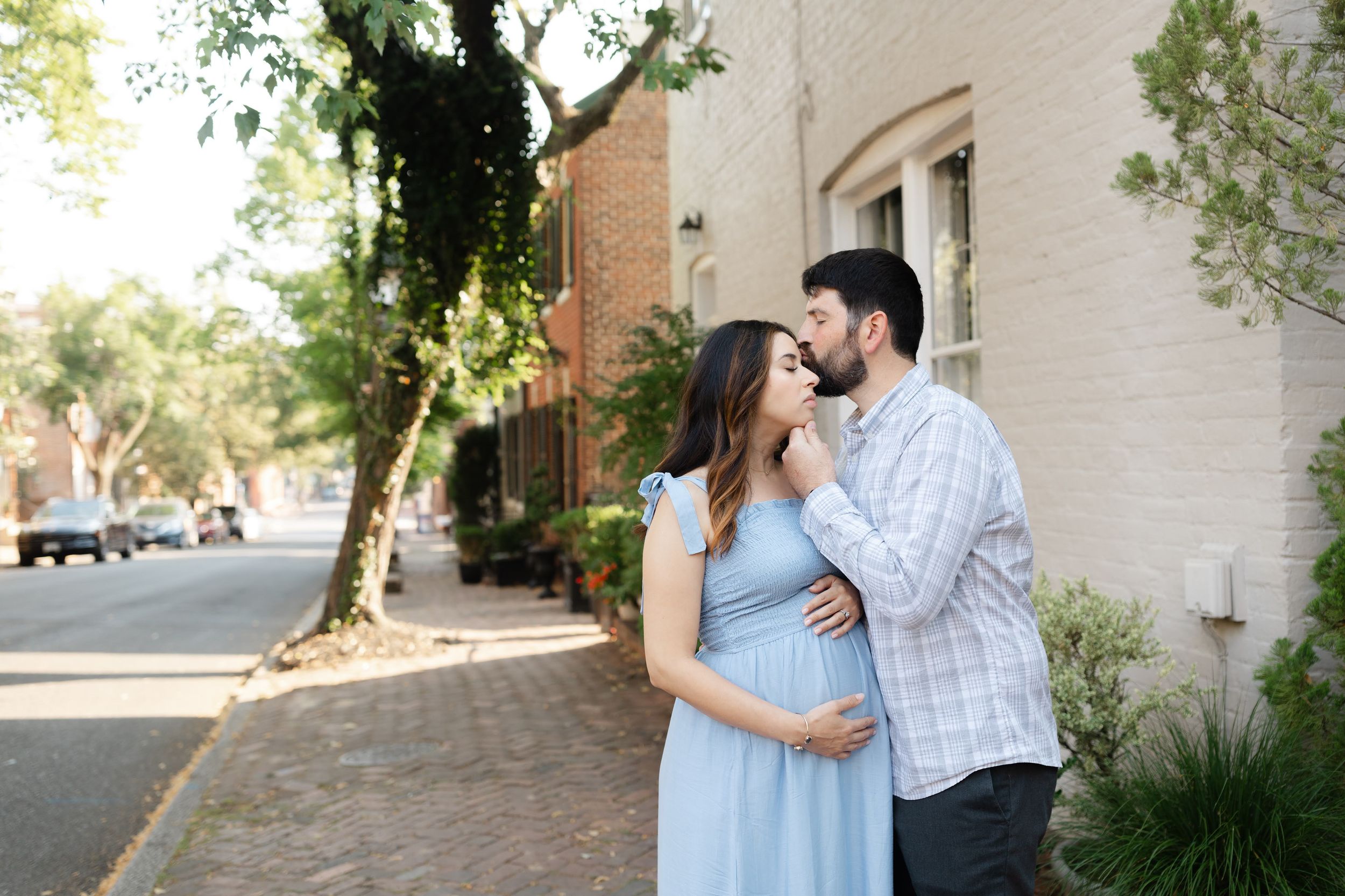 Pregnant couple embracing on a brick sidewalk, man kissing woman's head, she wears a light blue maternity dress.