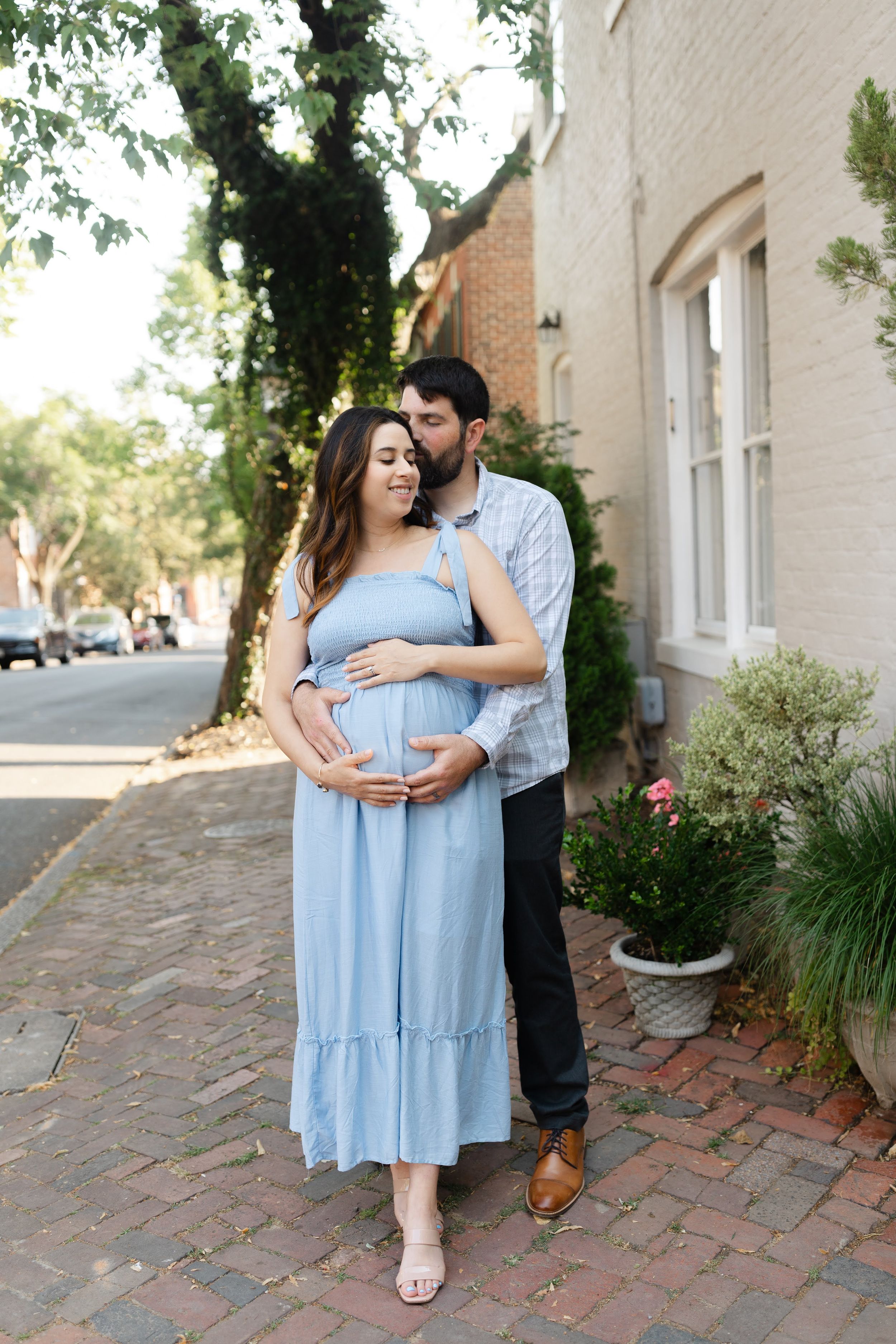 A couple shares a tender moment on a brick sidewalk, the woman in a blue maternity dress cradling her baby bump.