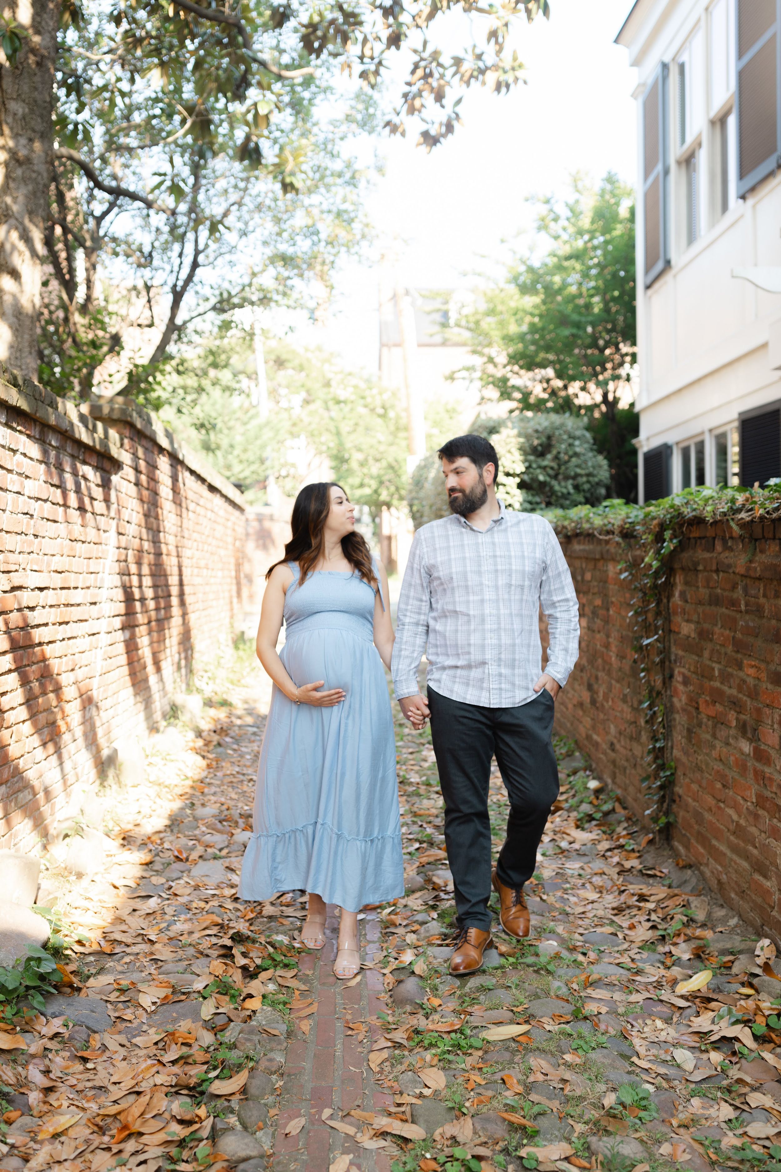 Pregnant woman in blue dress holds hands with bearded man walking along brick-walled path covered in autumn leaves.