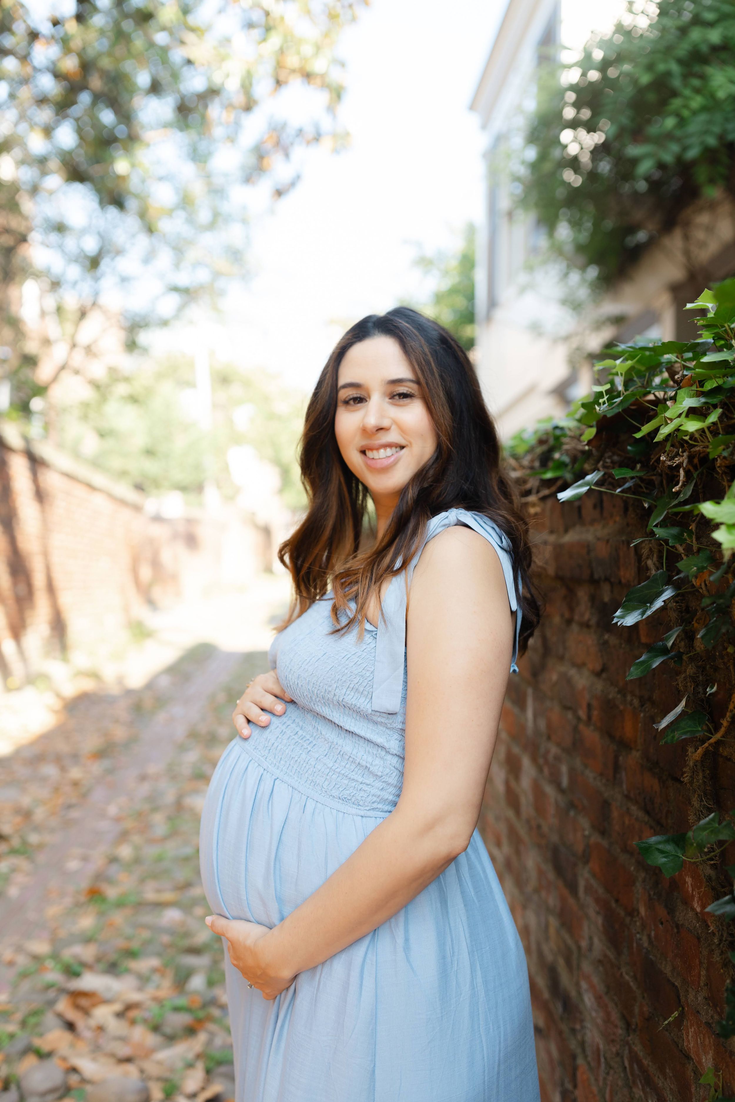 Smiling pregnant woman in blue dress cradling baby bump beside brick wall in outdoor alley setting.