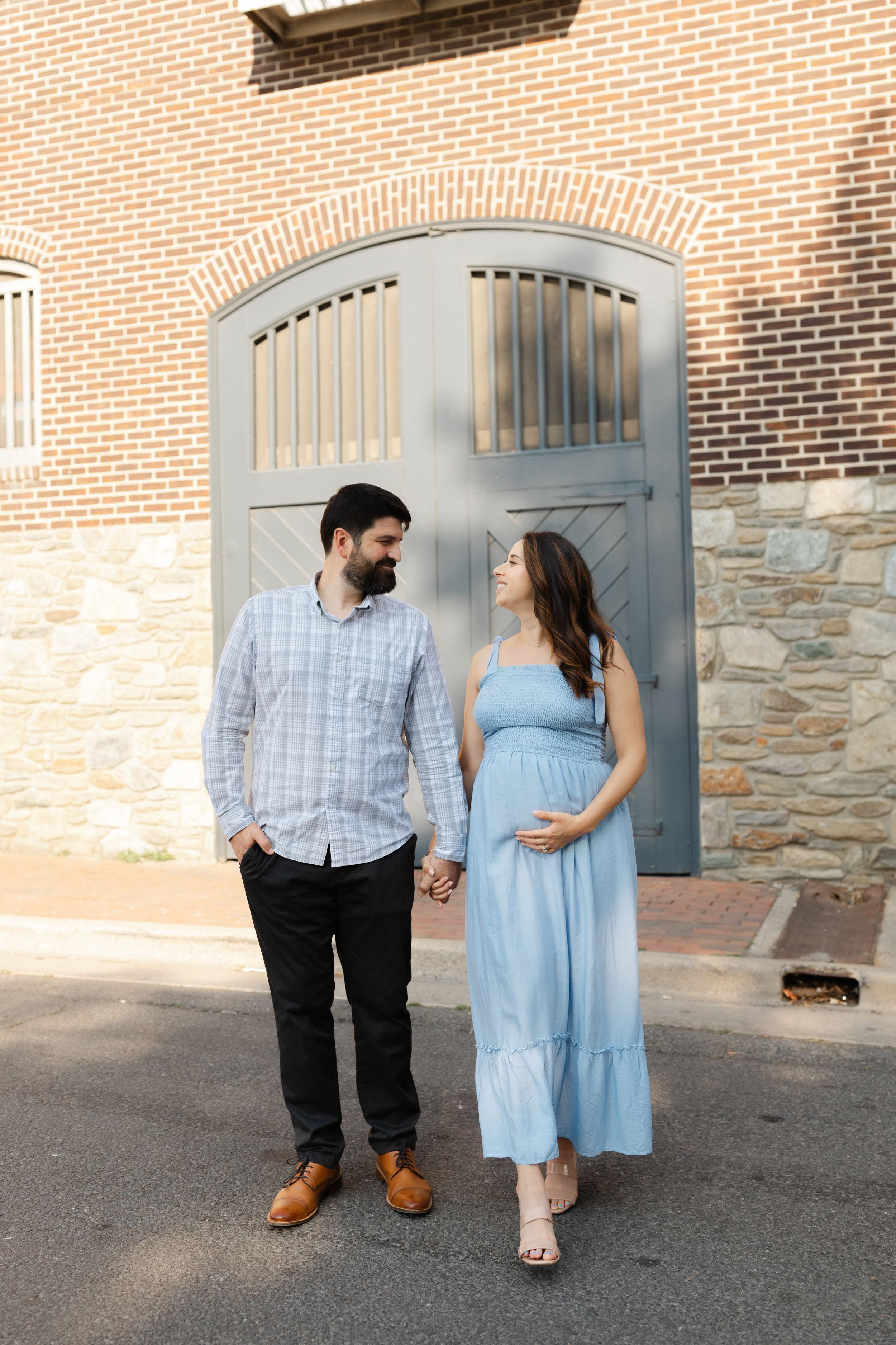 Pregnant couple holding hands in front of a blue arched door, woman in light blue maxi dress cradling baby bump.