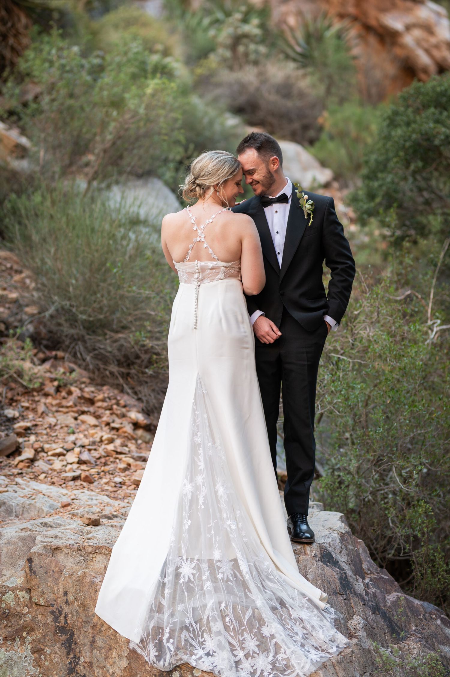 Bride in lace wedding gown and groom in black tuxedo share intimate moment on rocky path surrounded by desert greenery.