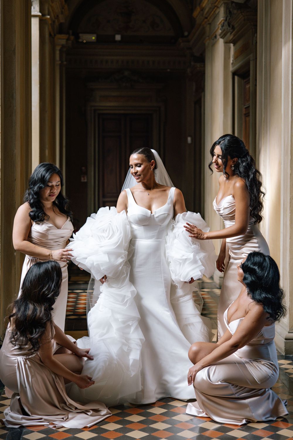Bride in white gown surrounded by bridesmaids in champagne dresses helping her prepare in elegant venue.