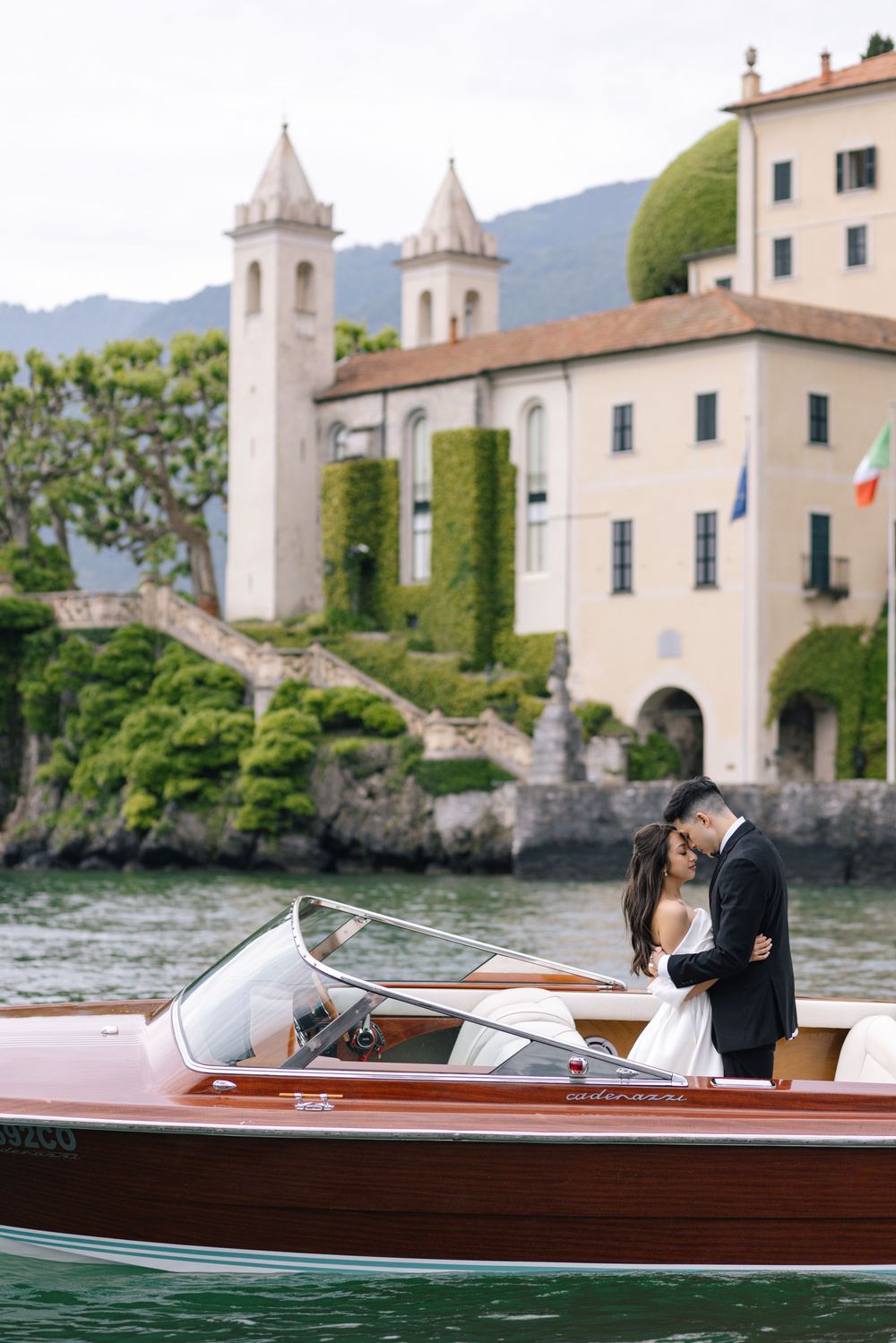 Couple embracing on a classic wooden boat on Lake Como, Italy, with an elegant ivy-covered villa in the background.