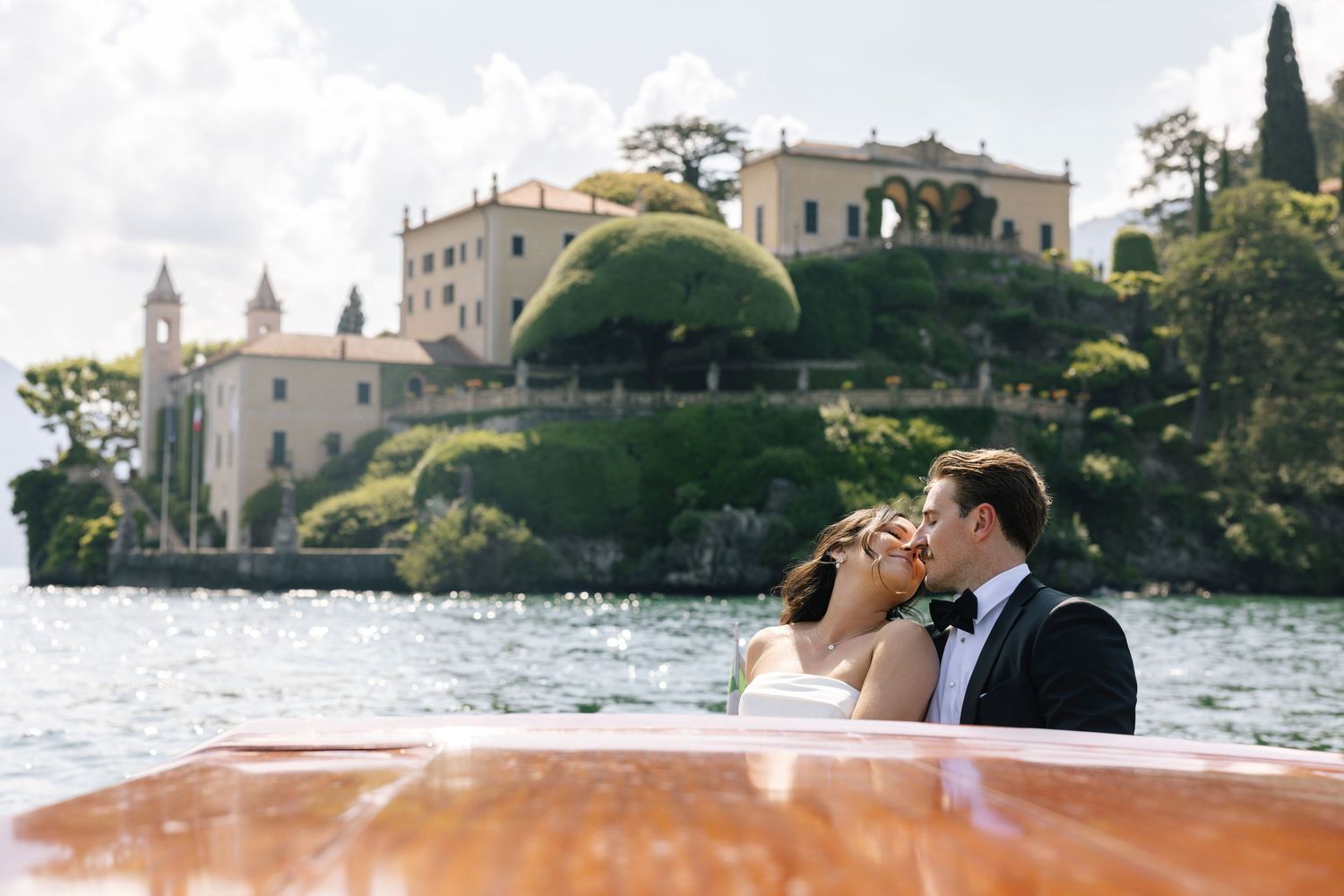 Couple sharing a romantic kiss on a wooden boat with Villa del Balbianello on Lake Como in the background.
