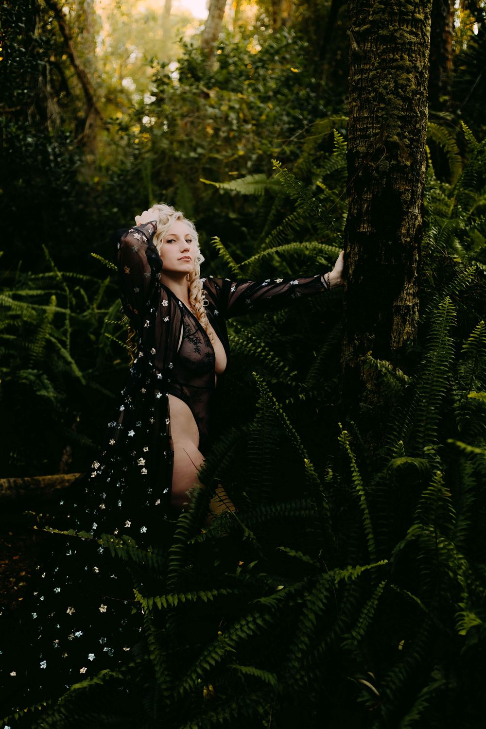 A blonde woman in a sheer black floral robe poses gracefully among lush green ferns in a forest setting.