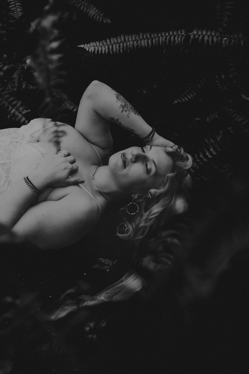 Black and white photo of a woman with tattoos lying among ferns, eyes closed, wearing a white dress and hoop earrings.