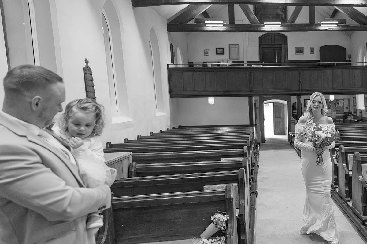 Black and white photo of a groom holding a child watching a bride walk down the church aisle on their Lake District elopement wedding day.