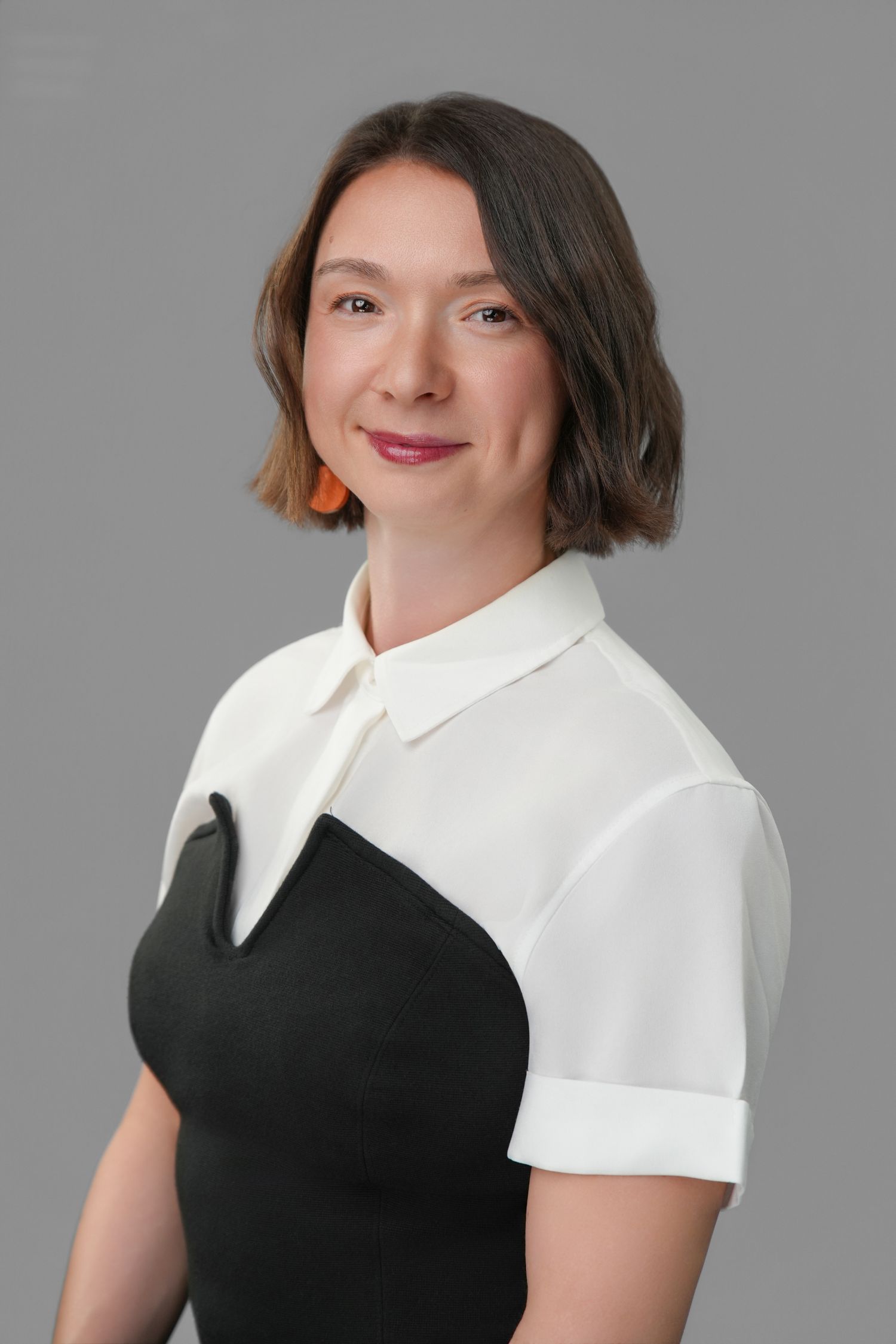 Professional woman in black dress with white collar shirt, short brown hair, orange earring, posing against gray background.