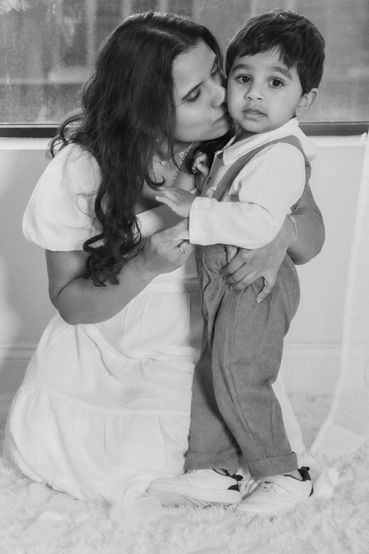 Black and white photo of a mother kissing her toddler son on the cheek while he looks at the camera.