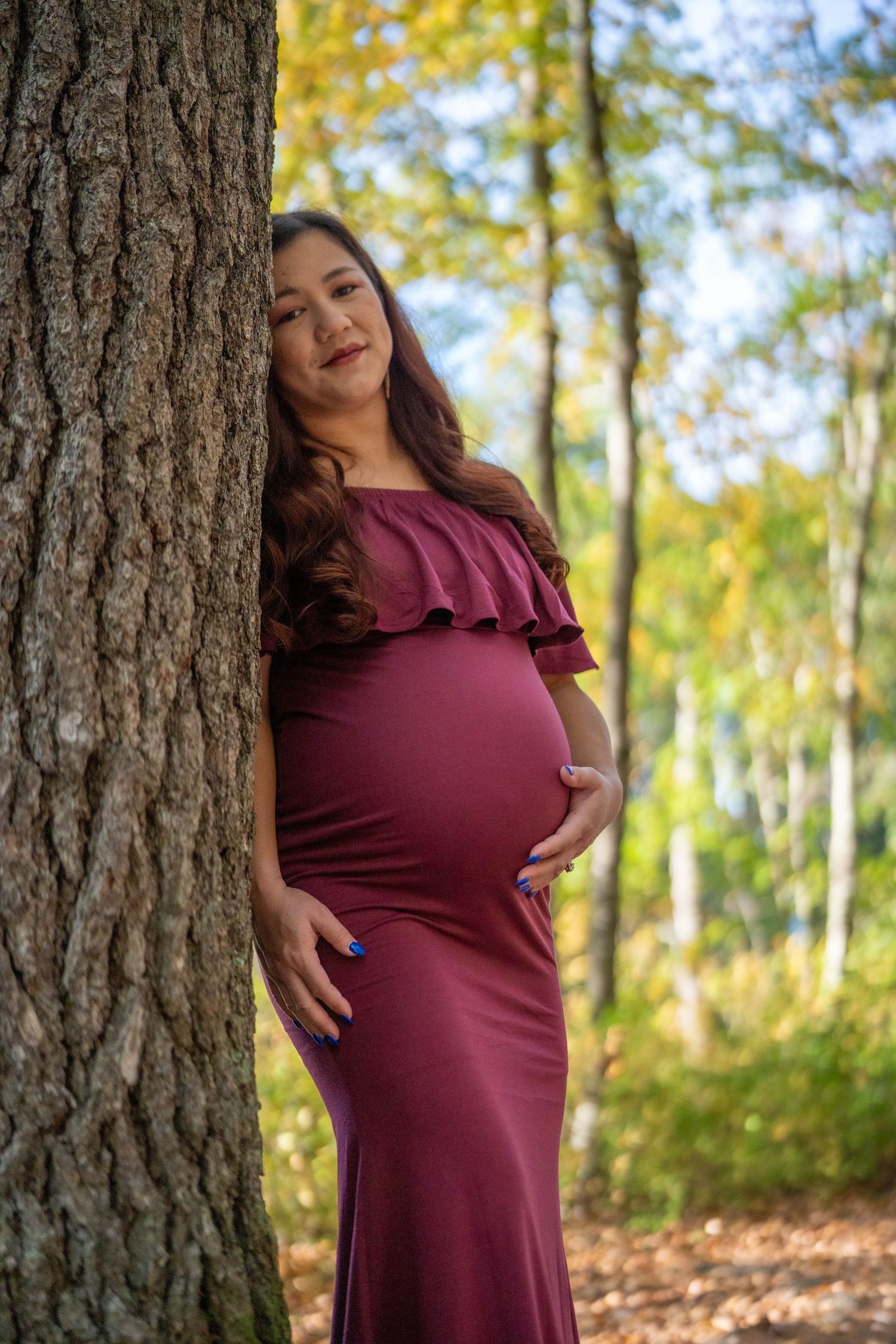 Pregnant woman in burgundy off-shoulder gown poses beside tree in autumn forest, cradling baby bump.