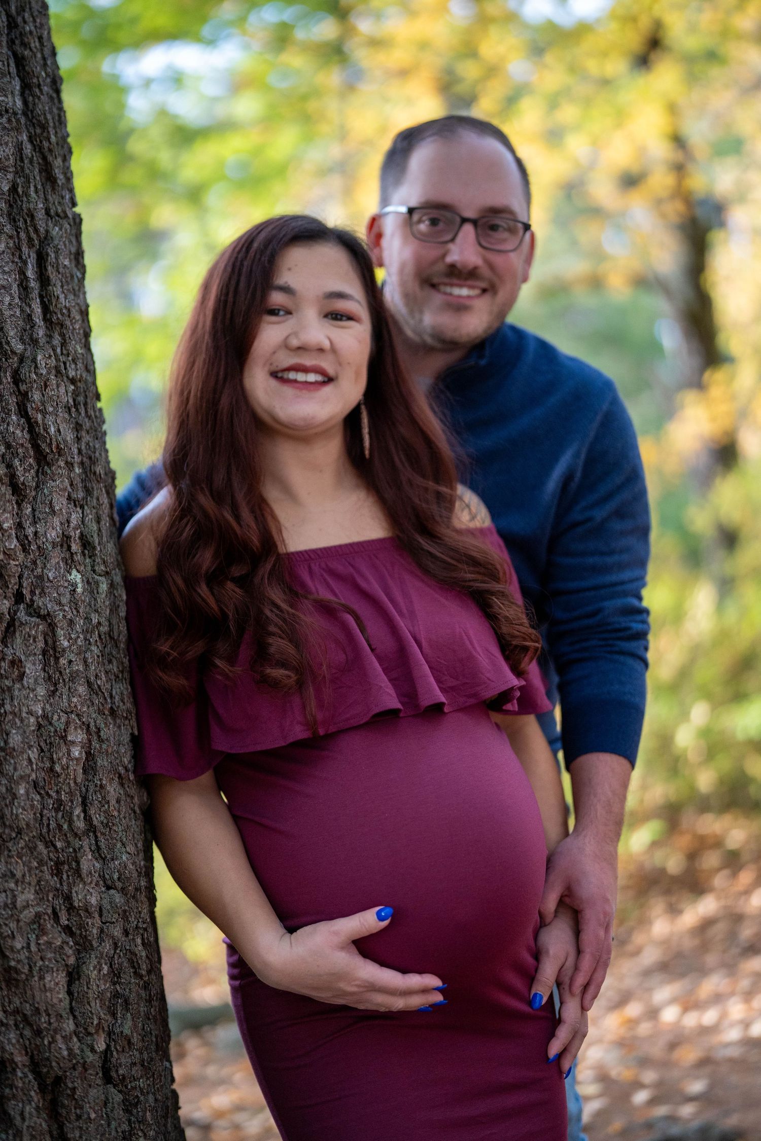 Pregnant woman in burgundy dress poses with man in blue sweater outdoors during fall season.