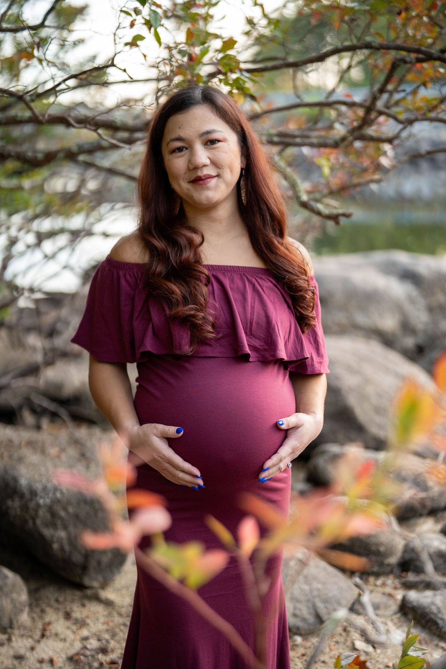 Pregnant woman in burgundy off-shoulder dress cradling baby bump outdoors among autumn foliage and rocks.