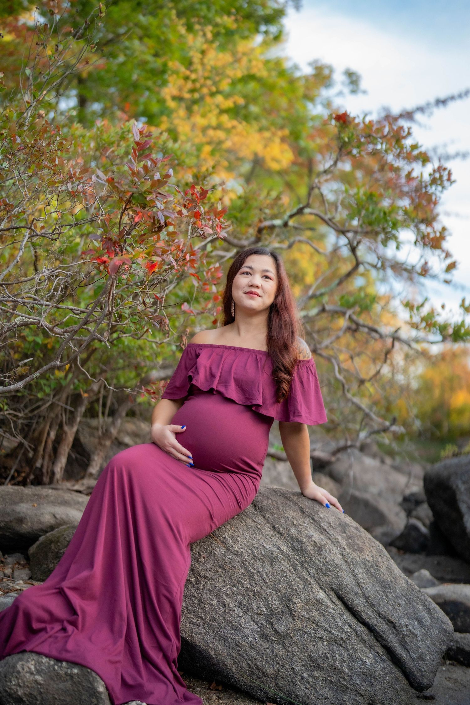 Pregnant woman in burgundy off-shoulder gown sitting on rocks surrounded by colorful autumn foliage.