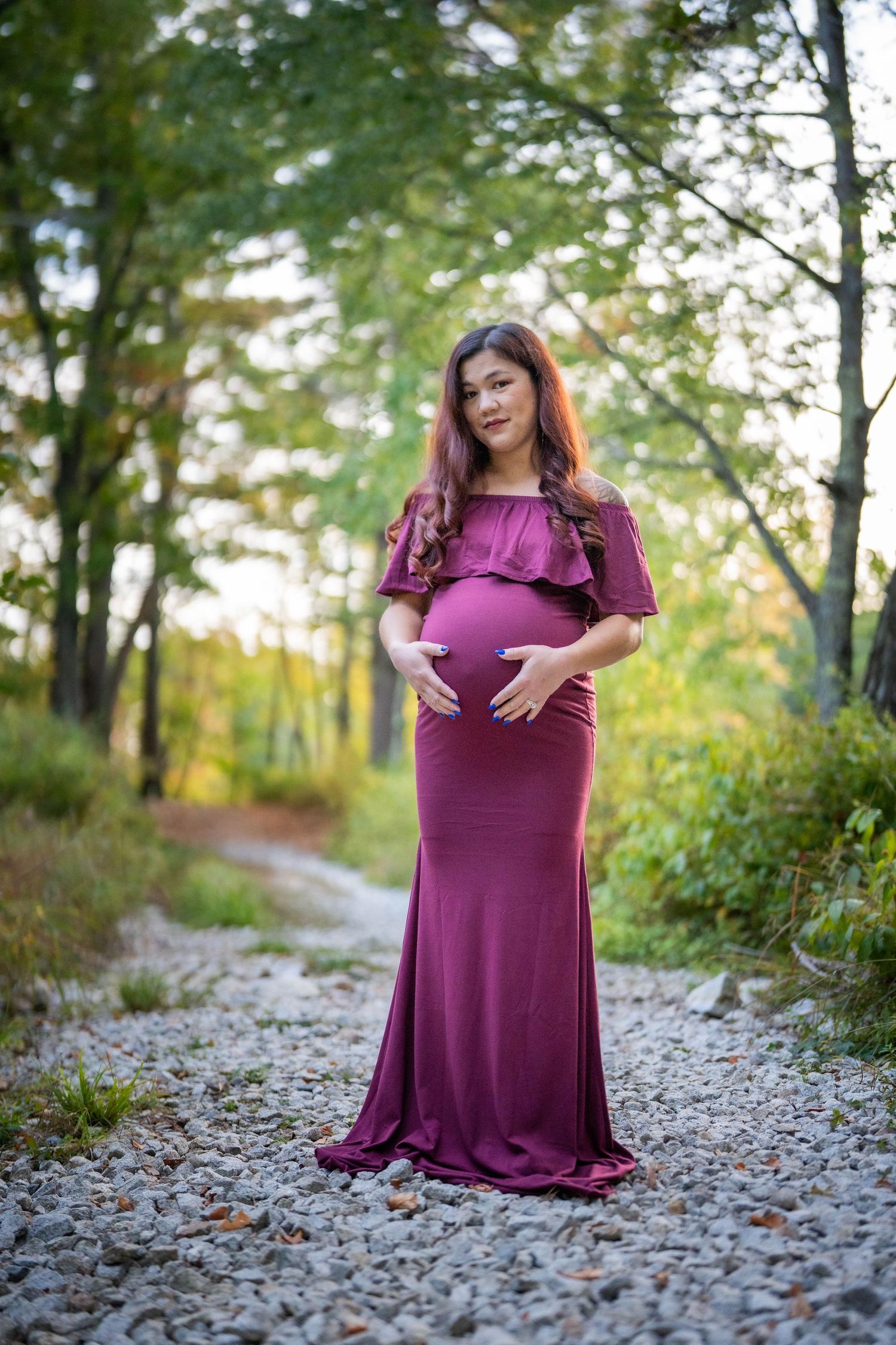 Pregnant woman in burgundy off-shoulder gown cradling baby bump on a gravel path surrounded by lush green trees.