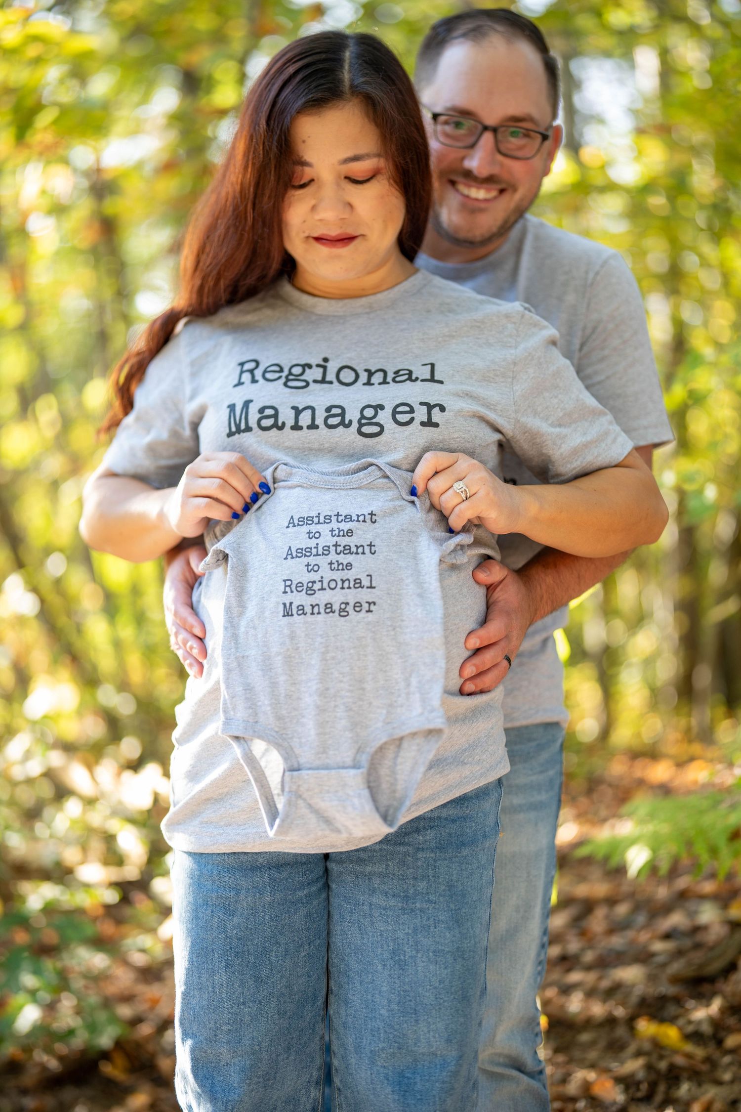 Couple holding baby onesie reading Assistant to the Regional Manager, woman wearing matching Regional Manager shirt.