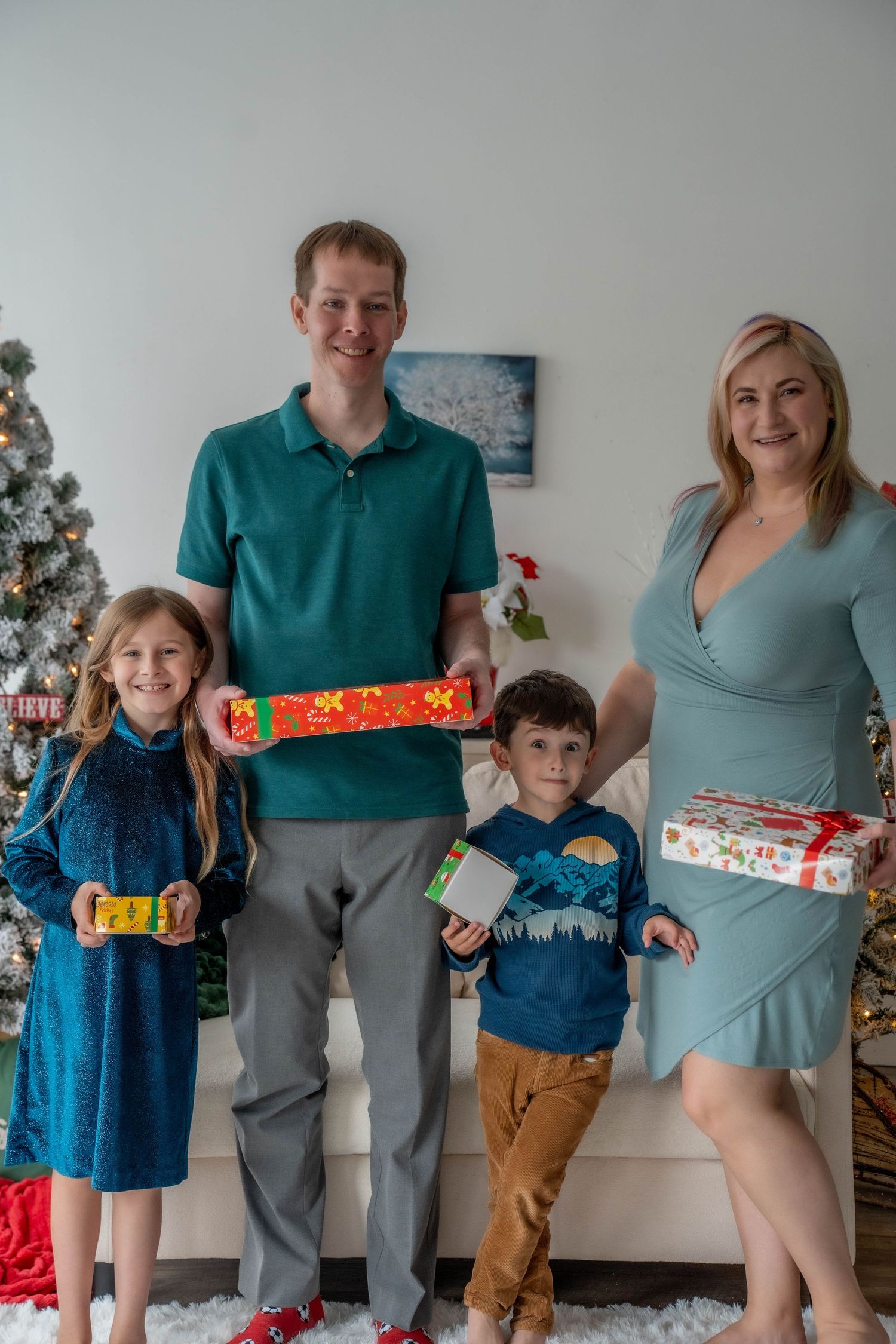 Family of four smiling and holding colorful Christmas gifts near decorated holiday trees in a festive living room.