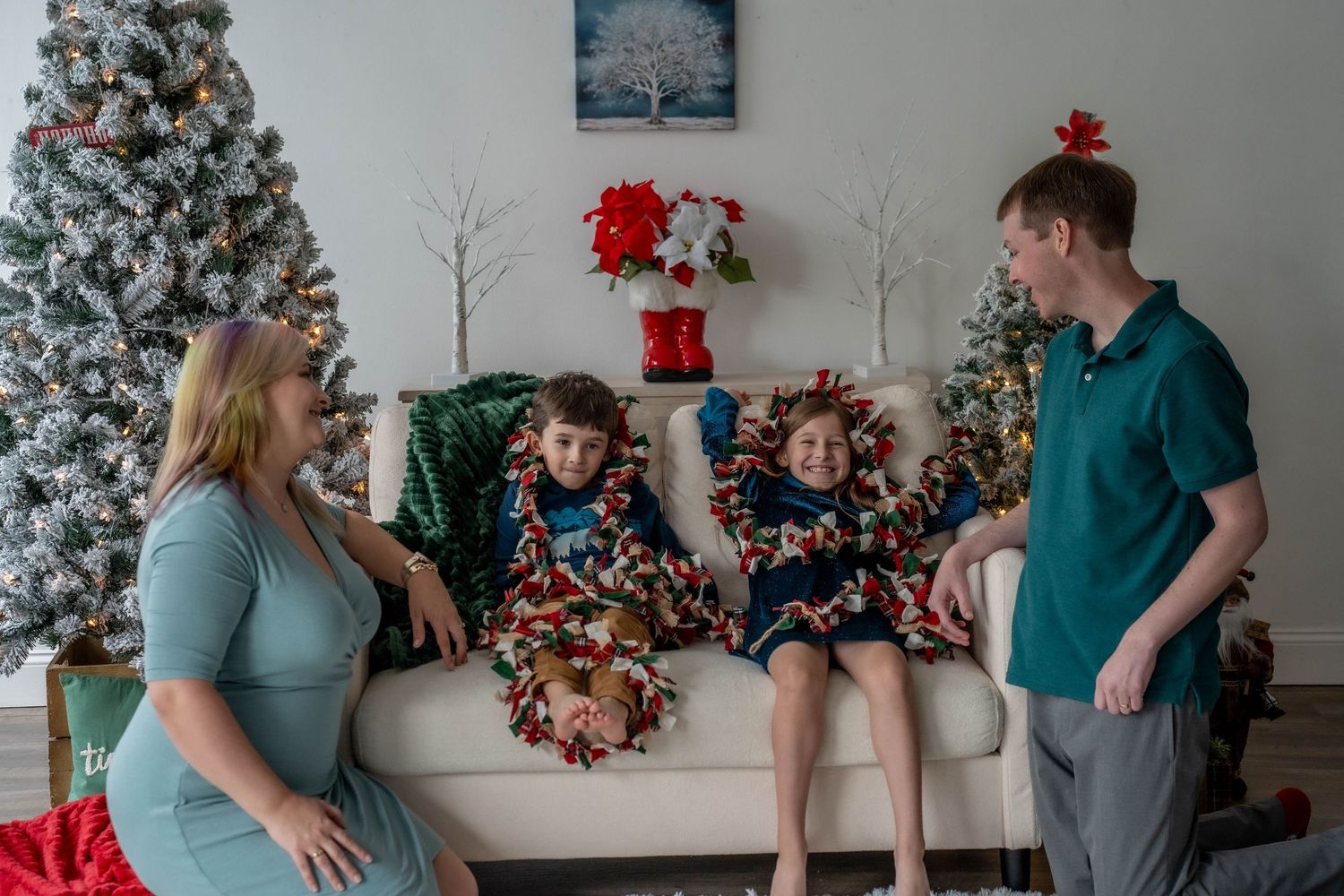 Family of four smiling together on a white couch surrounded by Christmas trees in a festive holiday living room.