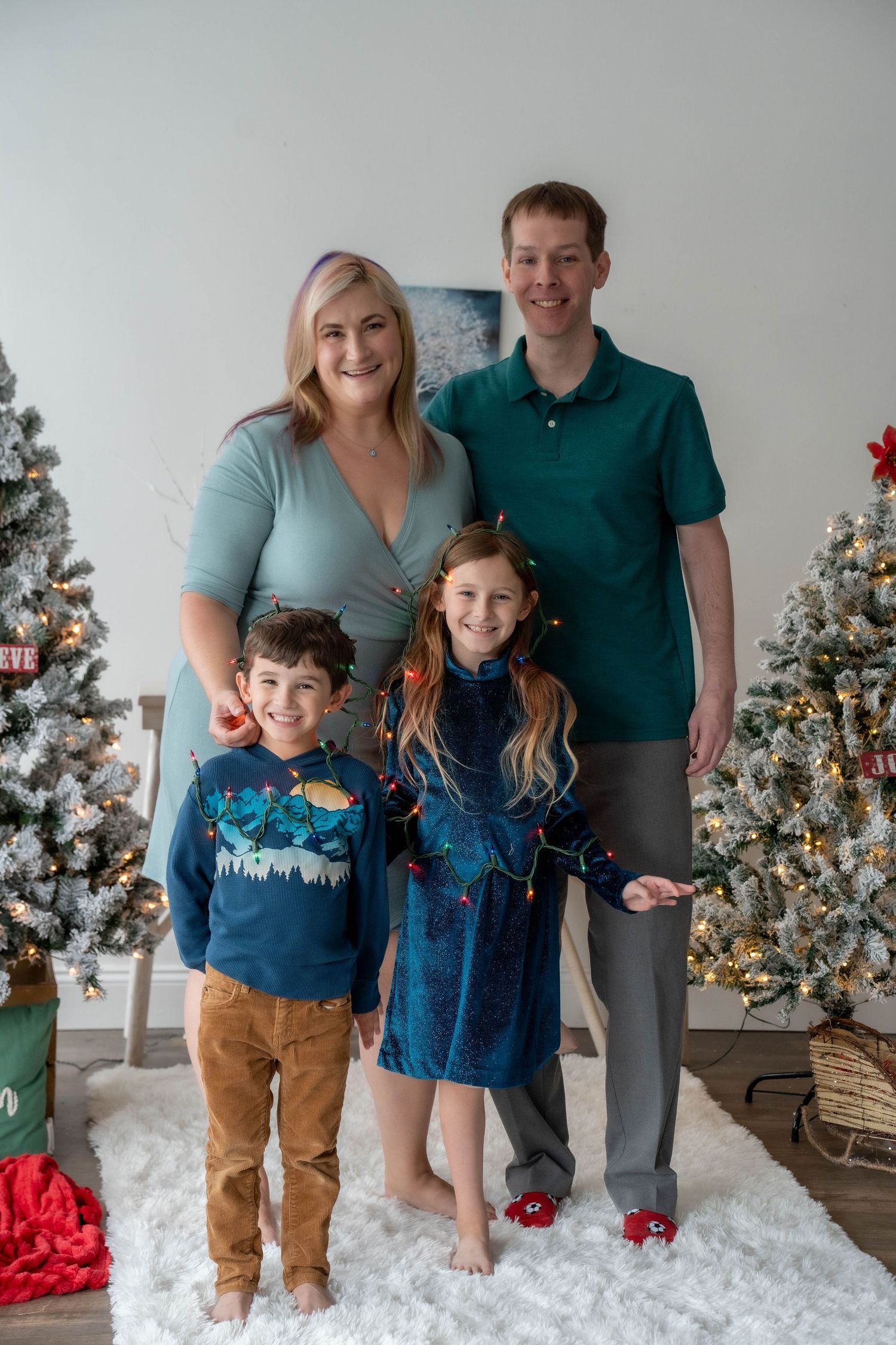 Family of four posing in front of Christmas trees, wearing coordinating blue and teal outfits, with girl wrapped in string lights.