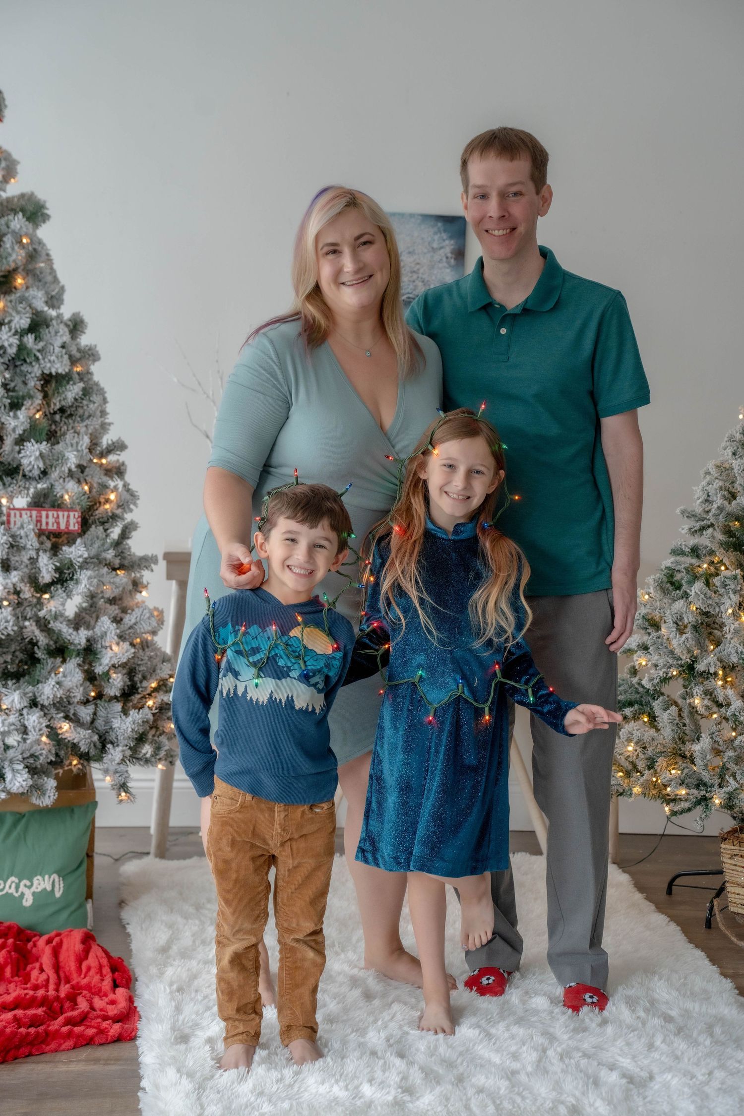 Family of four posing in front of Christmas trees, wearing coordinating teal and blue holiday outfits on a white fluffy rug.