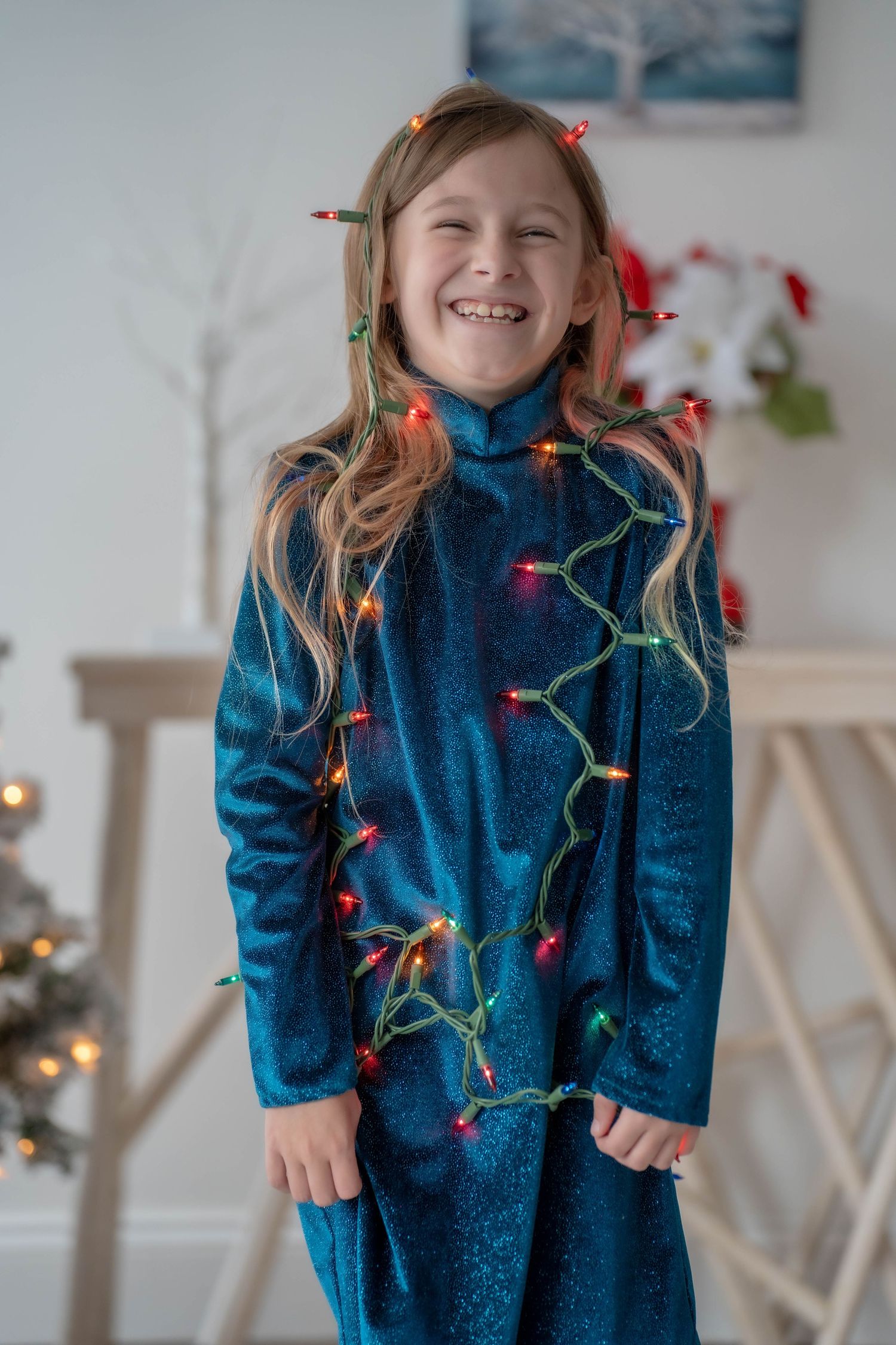Smiling girl in blue velvet dress wrapped in colorful Christmas lights, posing indoors near a decorated tree.