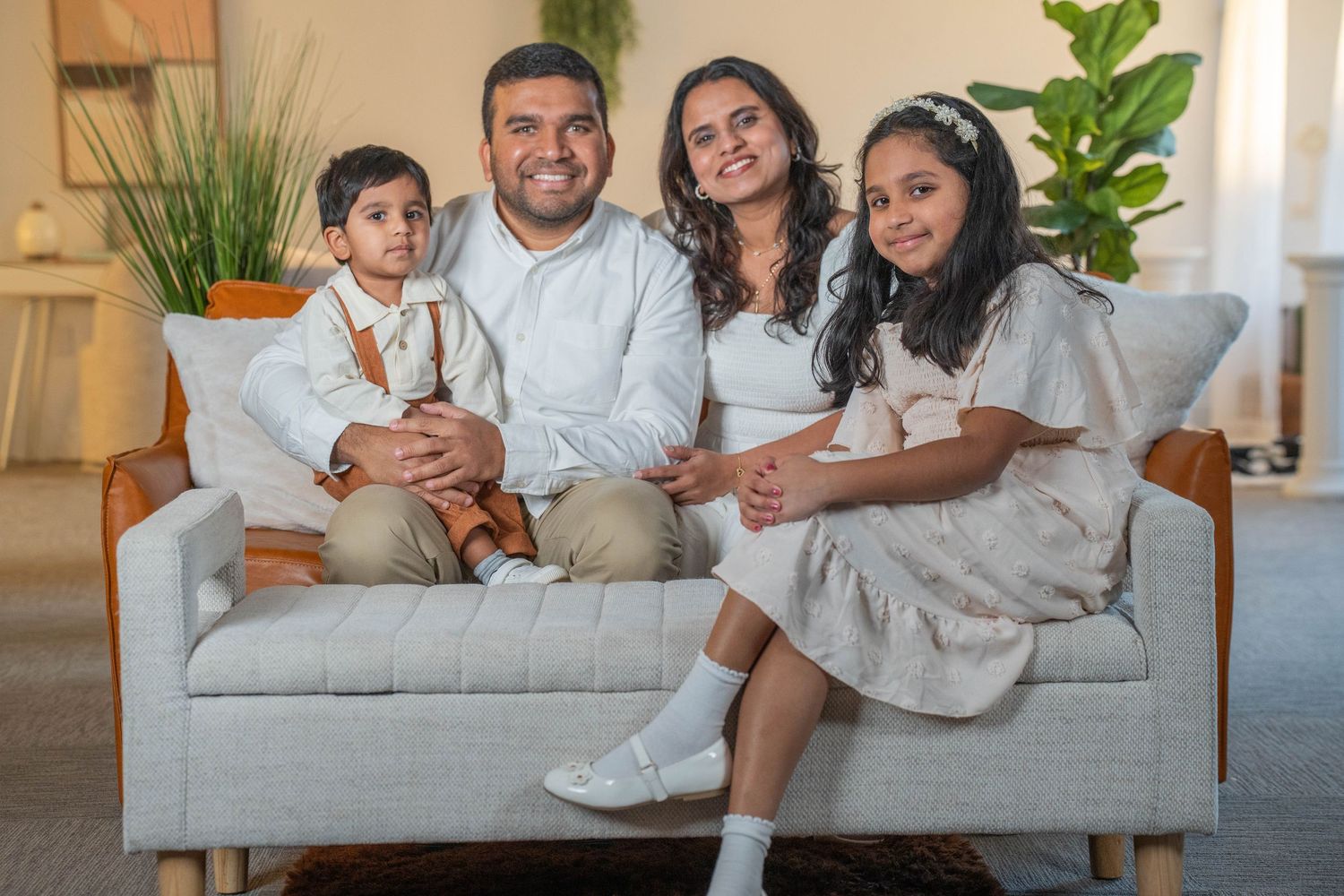 Happy South Asian family of four posing together on a gray sofa in a bright, modern living room.