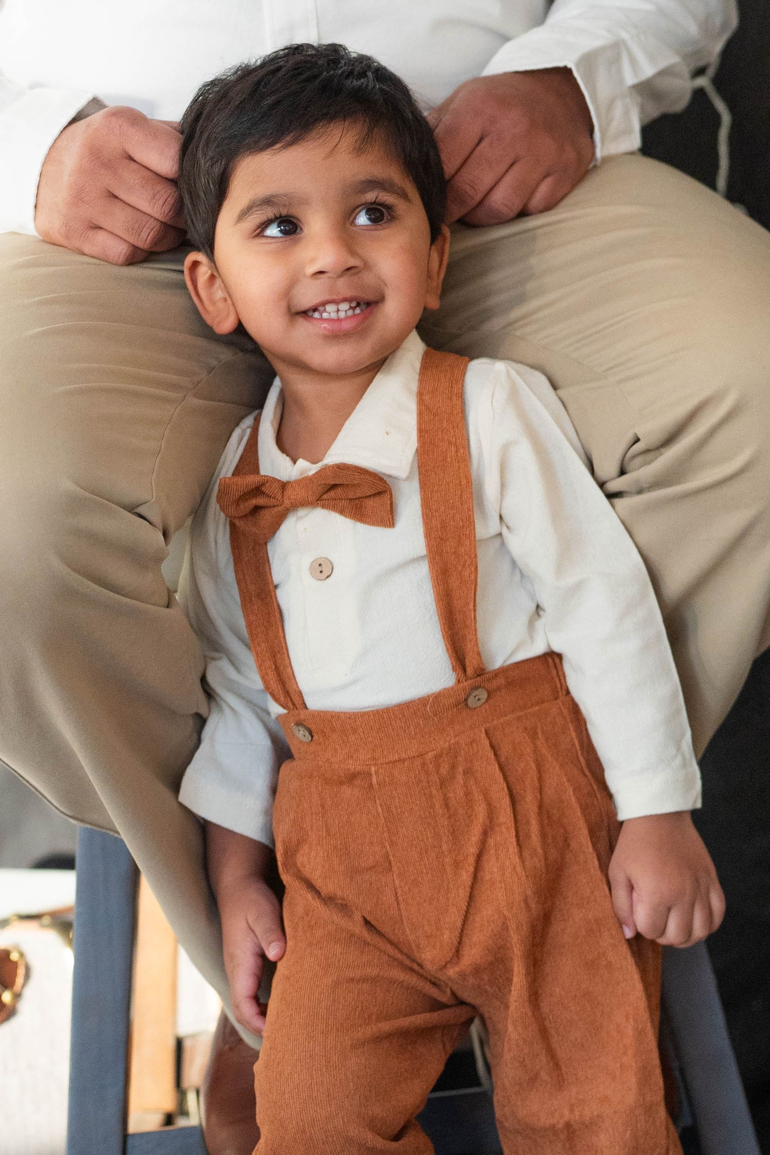 Adorable toddler boy in brown corduroy suspenders and bow tie smiling joyfully while standing in front of an adult.