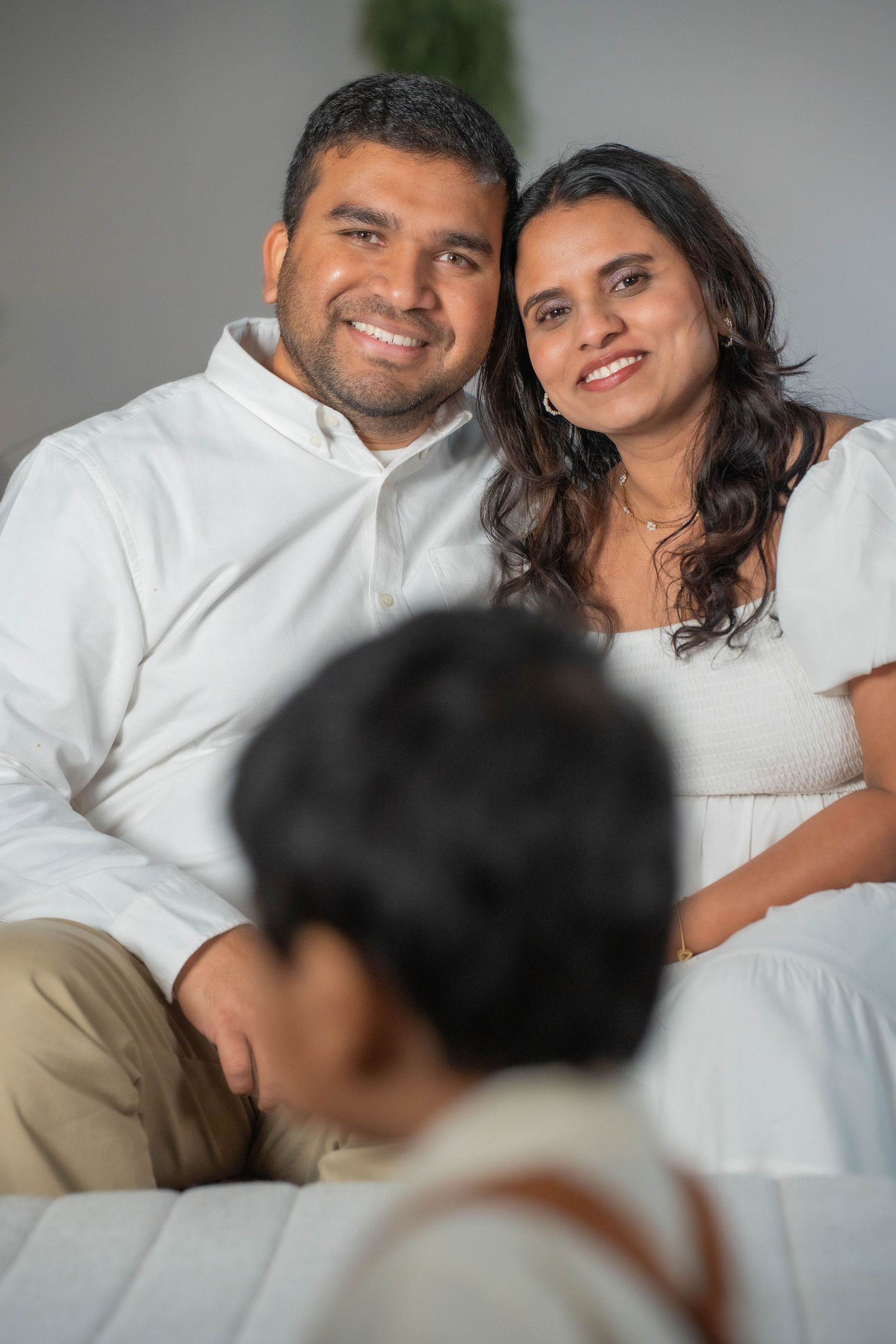 Smiling couple in white outfits sitting on a couch with a child in the foreground.