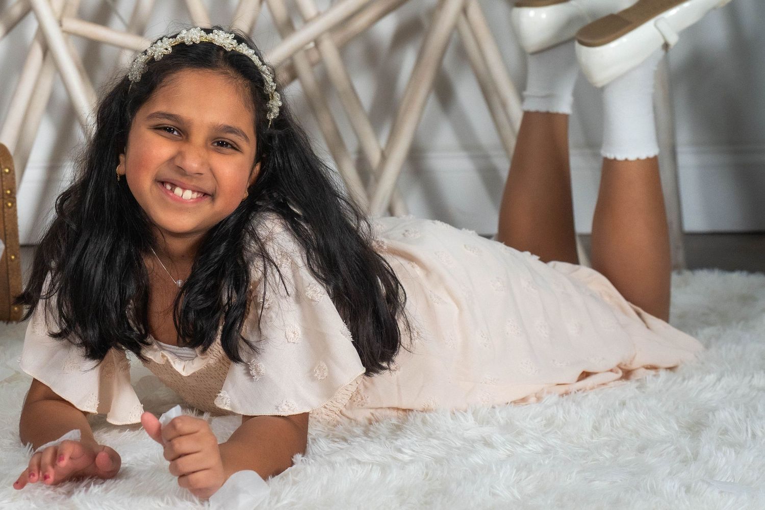 Young girl in white dress and floral headband smiling while lying on fluffy white rug.