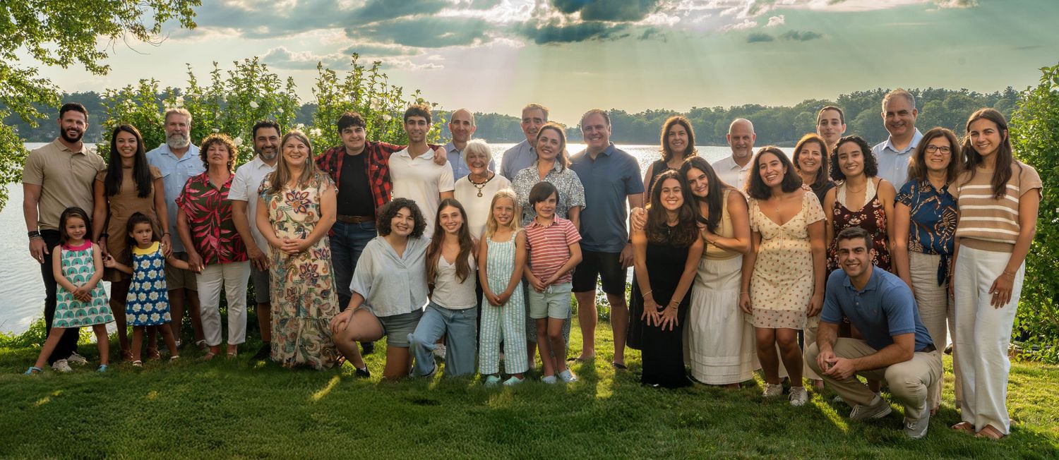 Large family group portrait outdoors on green lawn with scenic lake and dramatic sky in background.