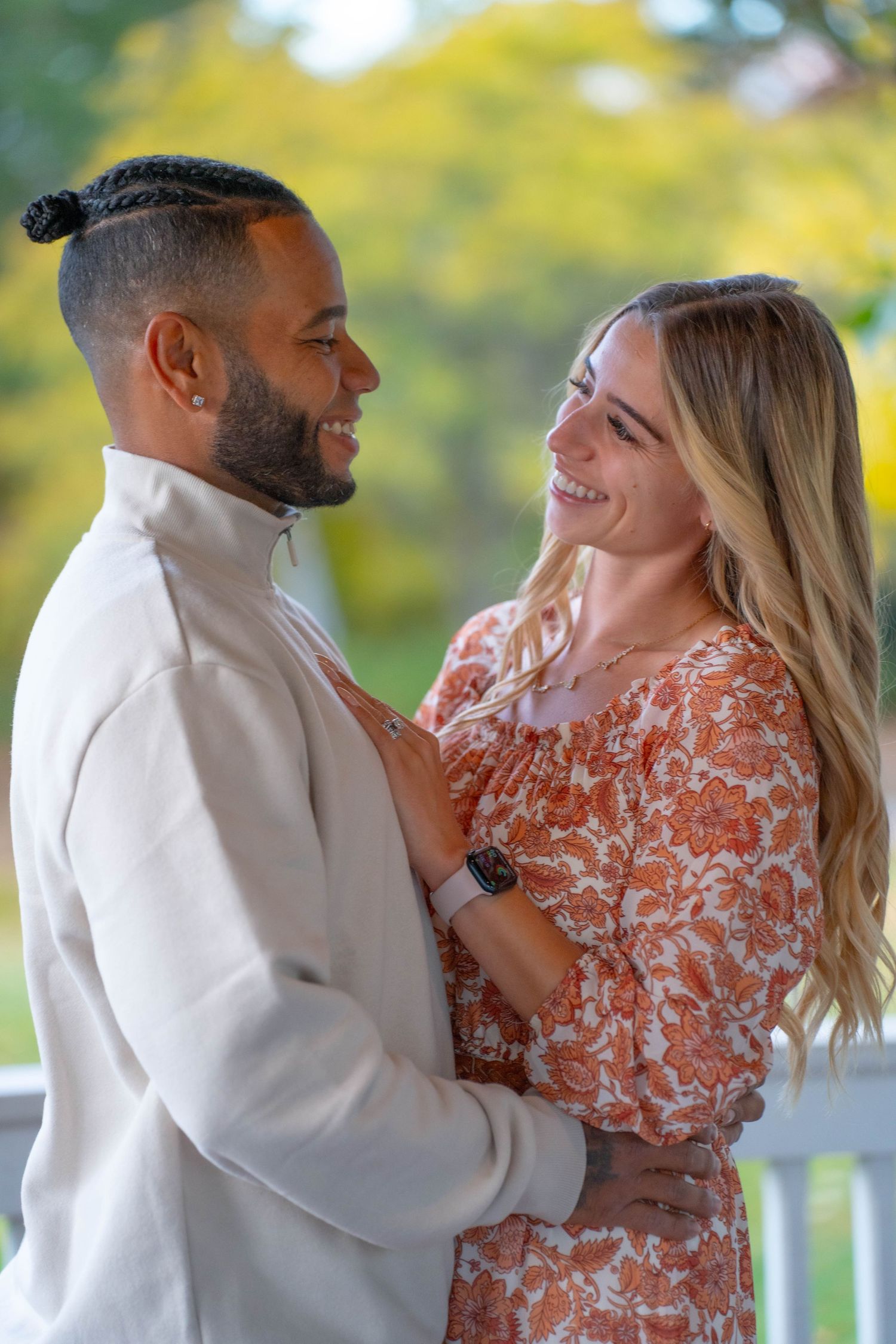 A smiling interracial couple embraces outdoors, woman in floral dress and man in white jacket, with autumn foliage background.