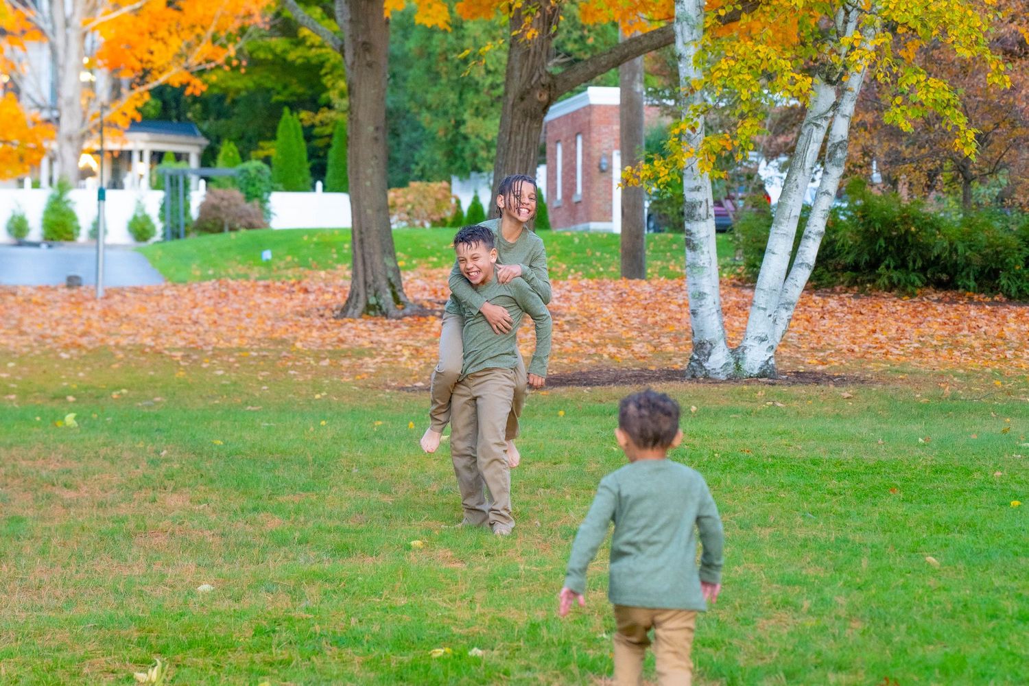 Family playing in autumn park, father giving child piggyback ride while another child runs toward them.