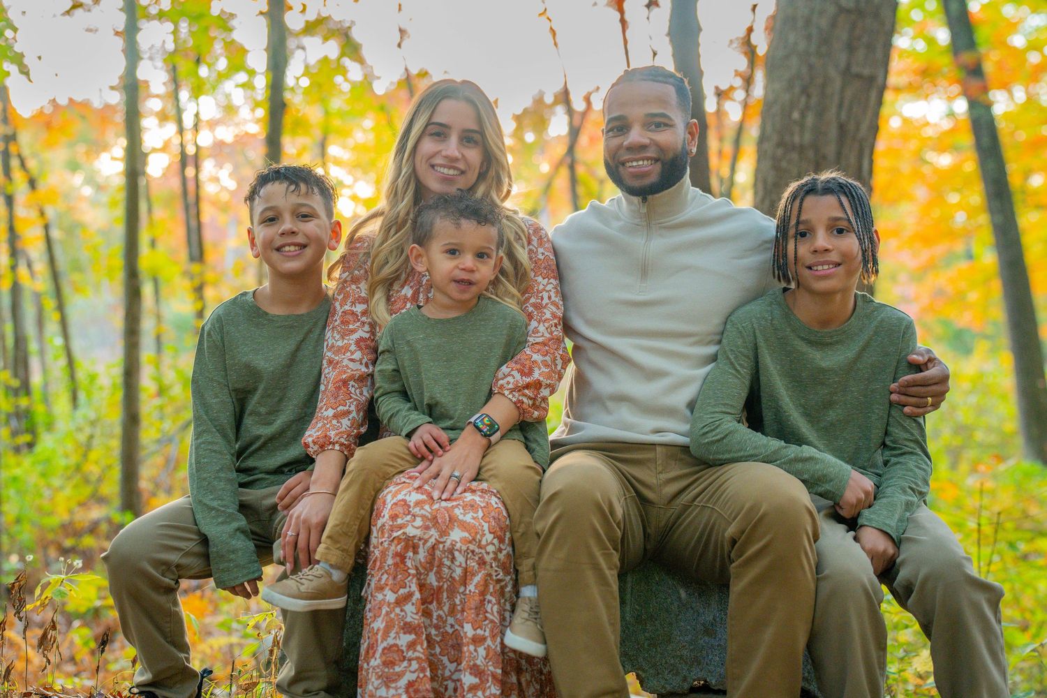 Family of five posing outdoors in autumn forest, wearing coordinated green and neutral outfits, smiling warmly together.