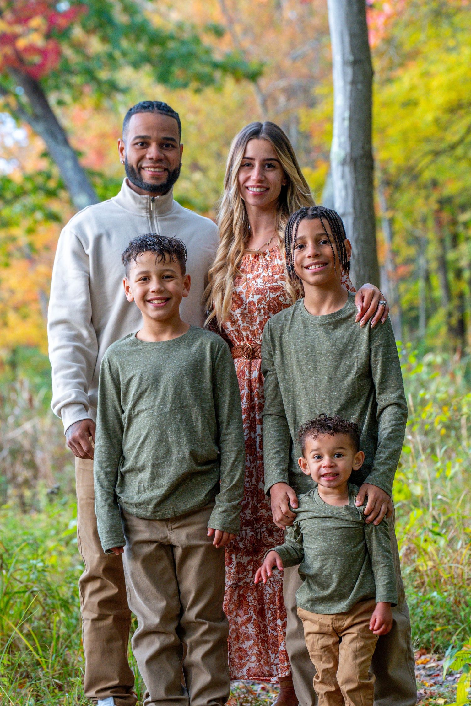 Family of five posing outdoors in fall foliage, wearing coordinating green and tan outfits, smiling at camera.