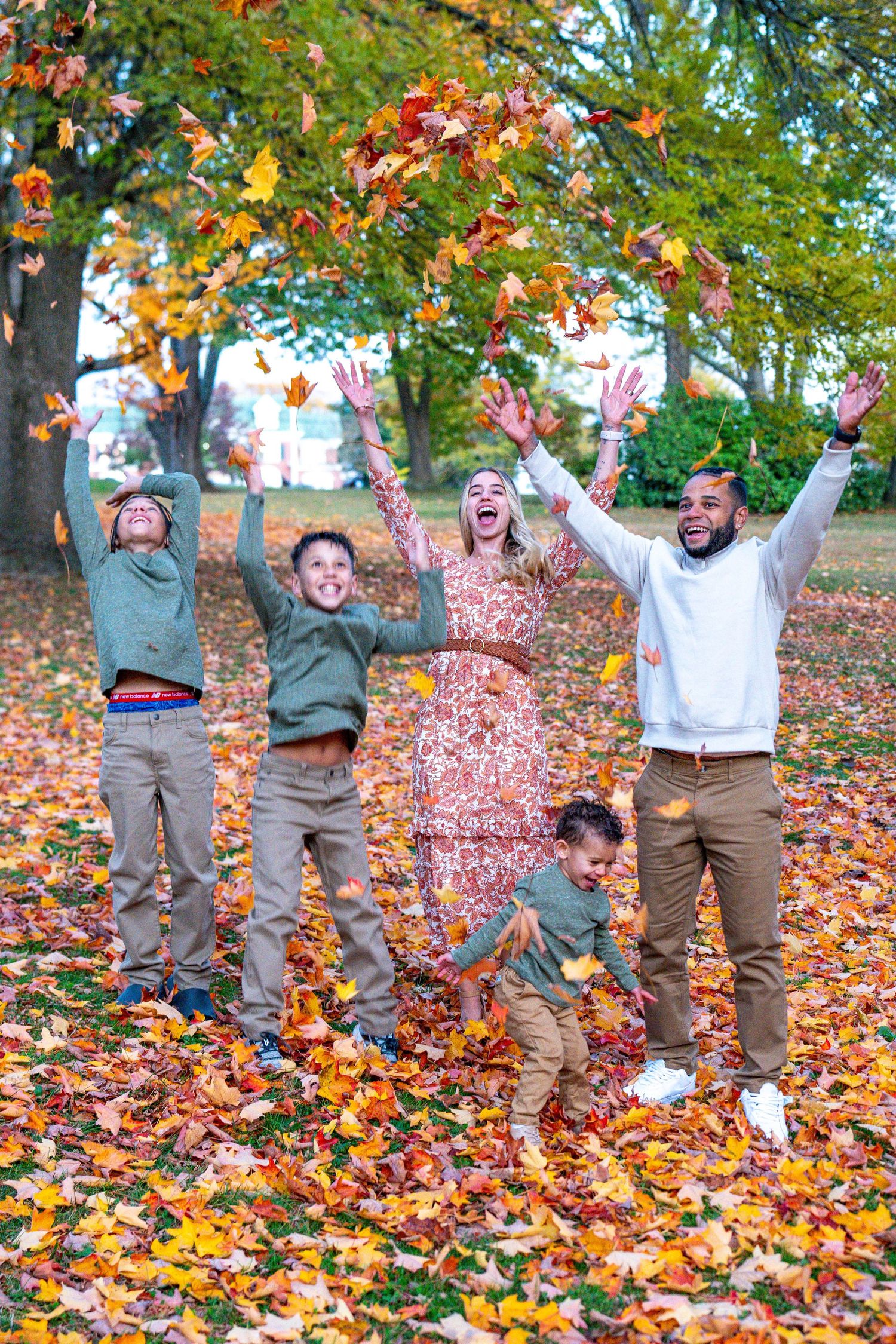 Family of five joyfully tossing colorful autumn leaves in a park, surrounded by vibrant fall foliage.