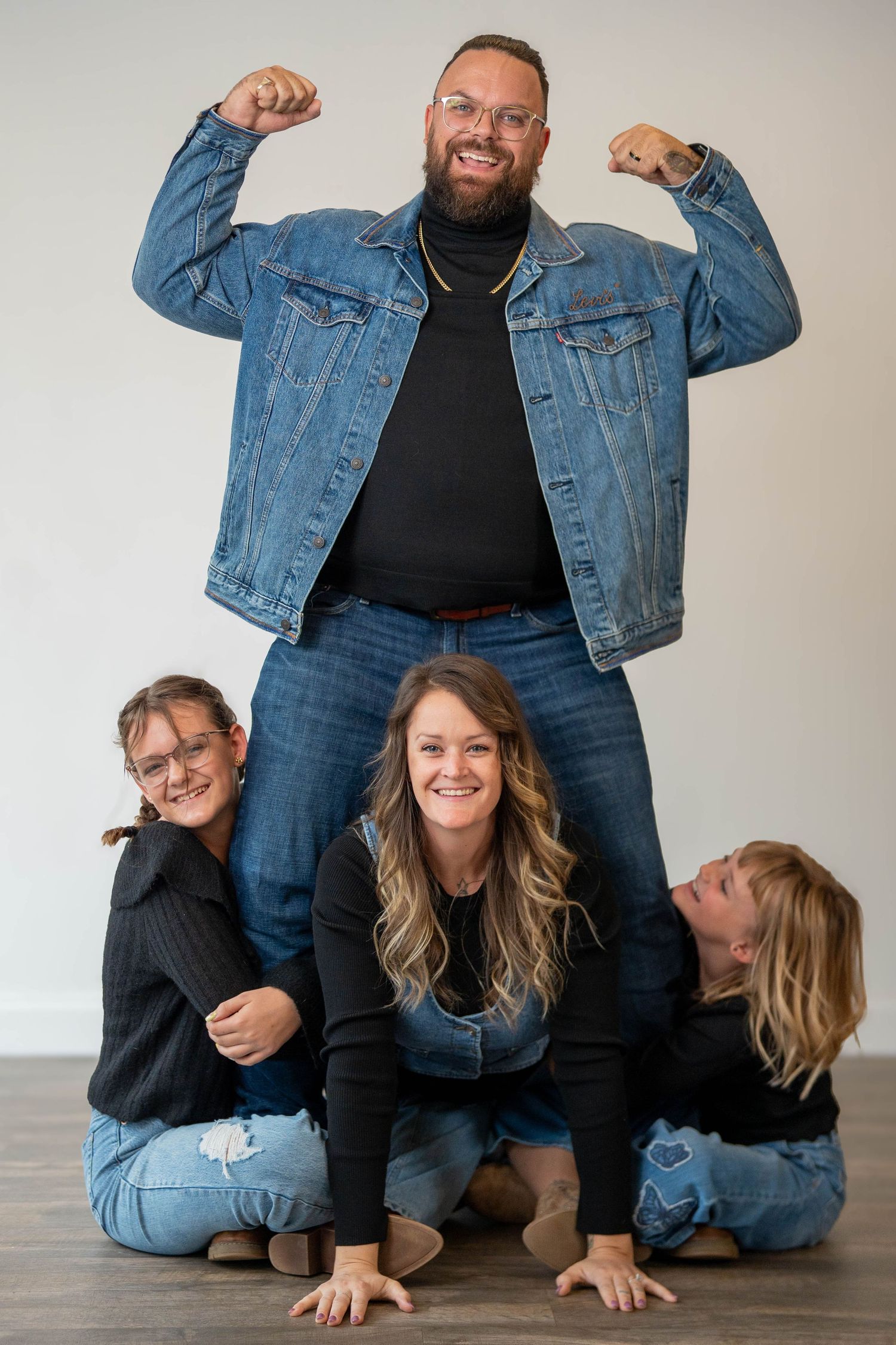 Family of four poses playfully, dad flexing above mom and two kids on floor, all wearing jeans and black tops.
