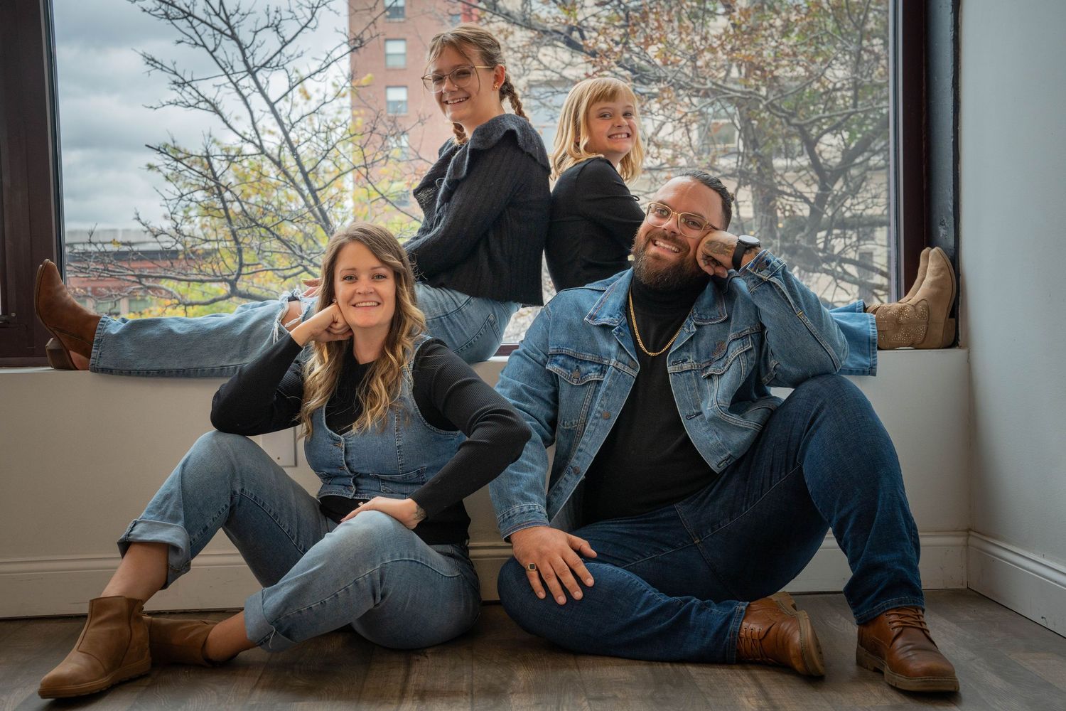 Four friends pose together near a large window, wearing denim outfits and brown boots in a bright indoor setting.