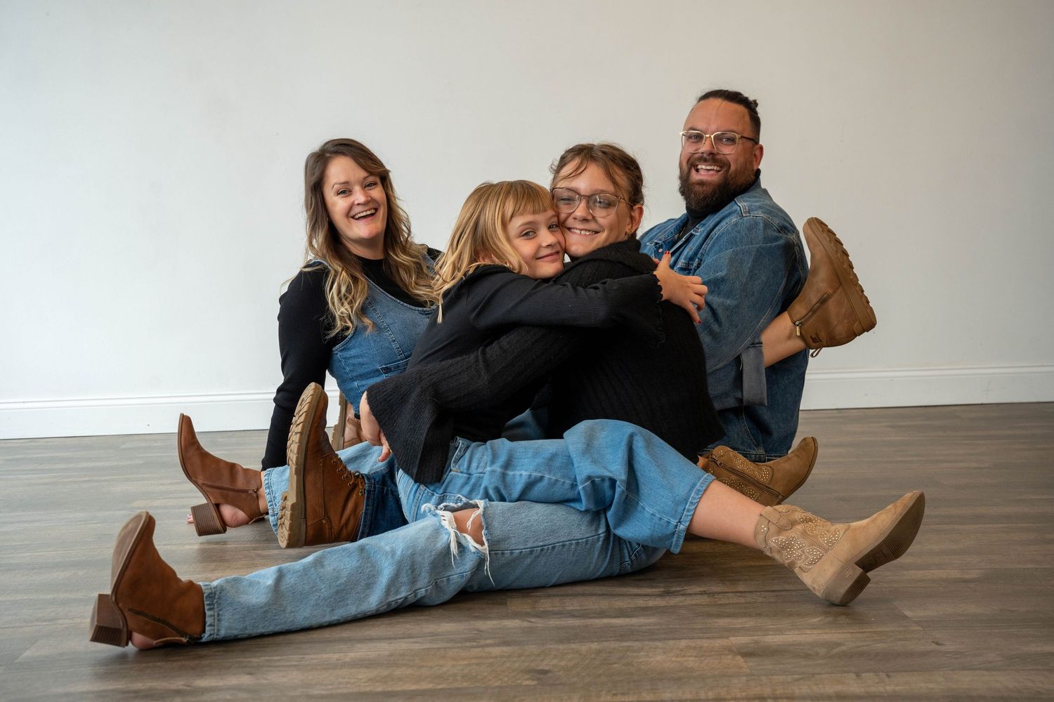 Four friends laughing and hugging on a hardwood floor, wearing jeans and boots in a bright white room.