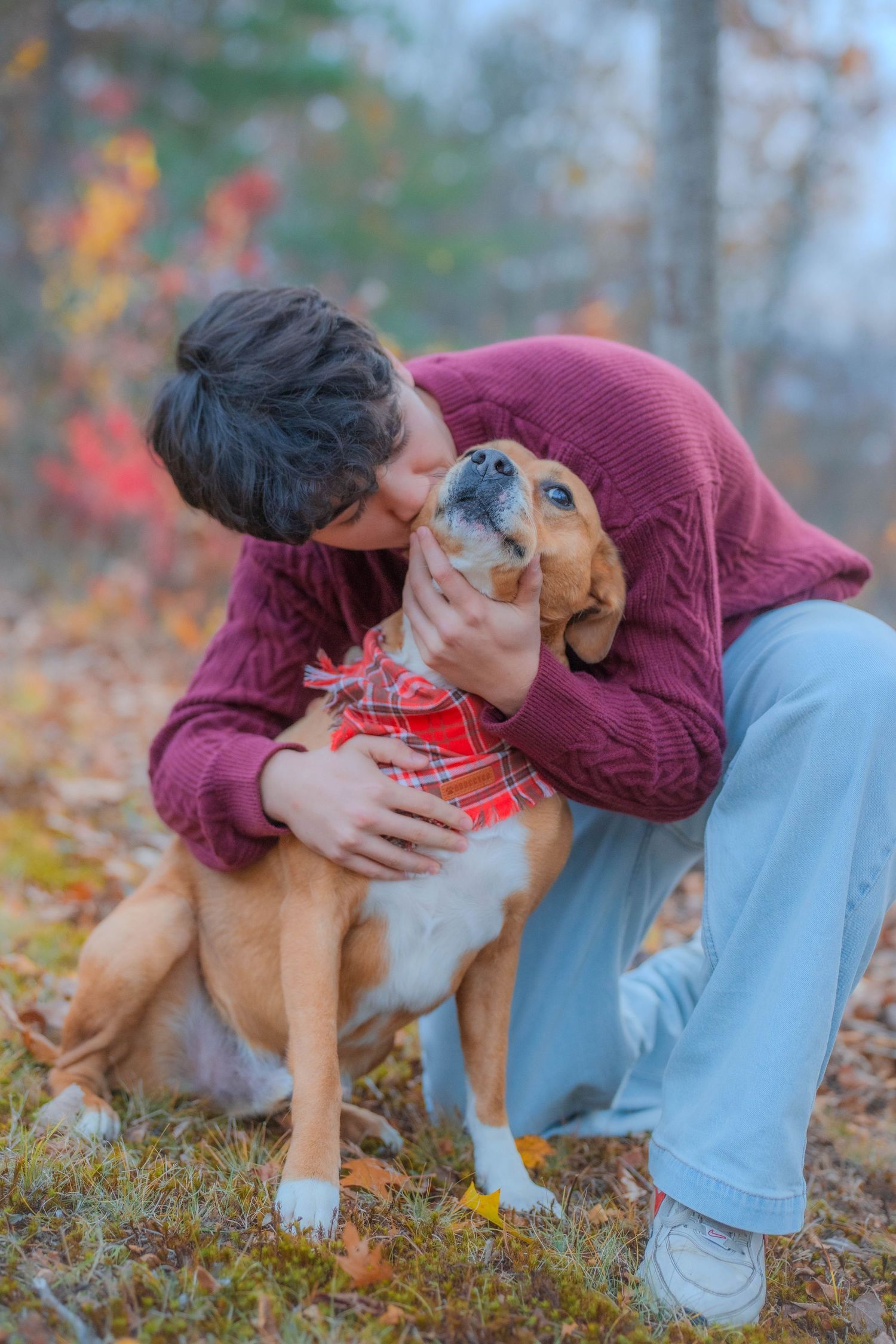 Person in maroon sweater kissing a beagle dog wearing a red plaid bandana in an autumn forest setting.