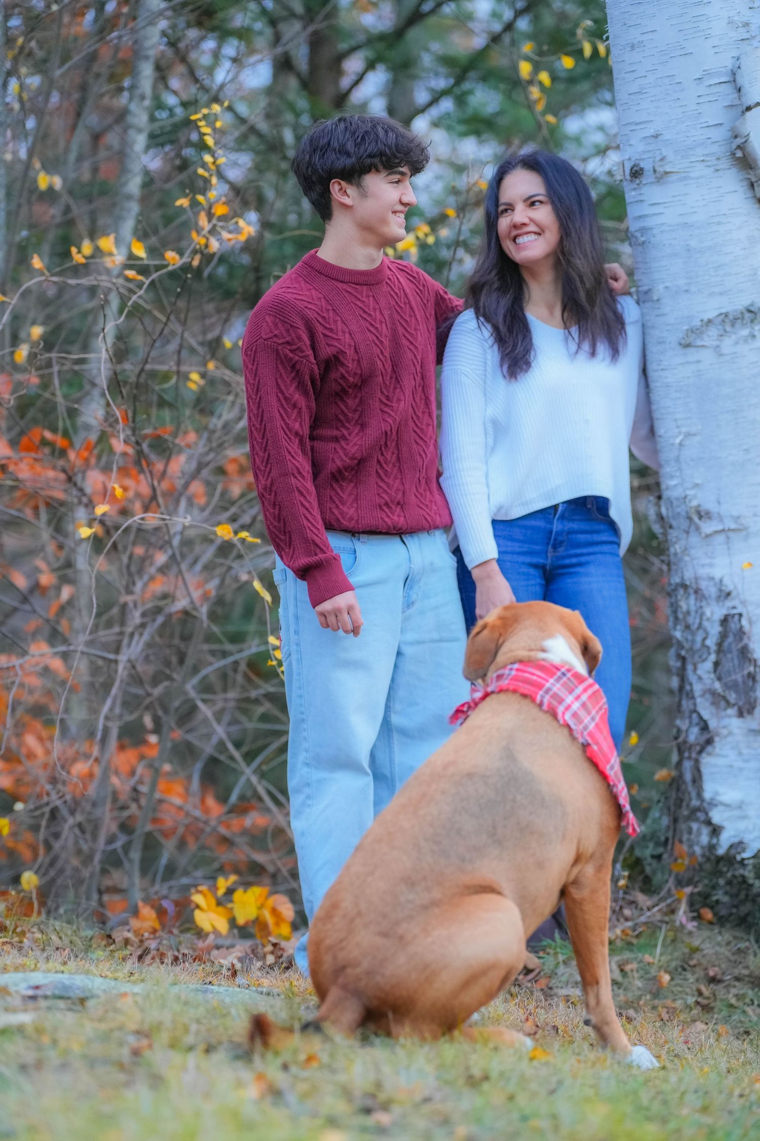 A young couple in fall sweaters and jeans smile at each other outdoors beside a birch tree with their brown dog wearing a plaid bandana.