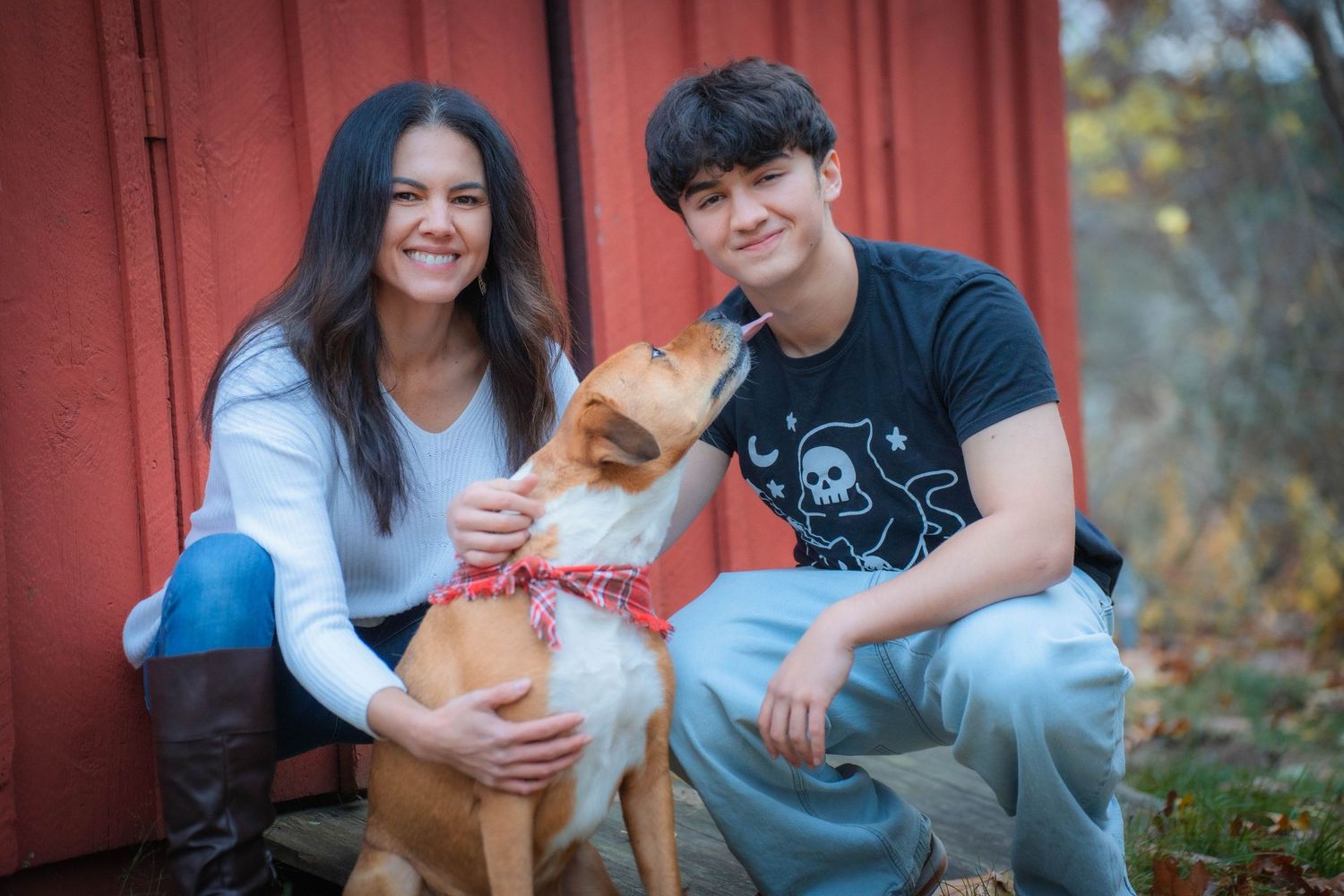 Two smiling young people pose with an adorable tan dog near a red barn wall outdoors.