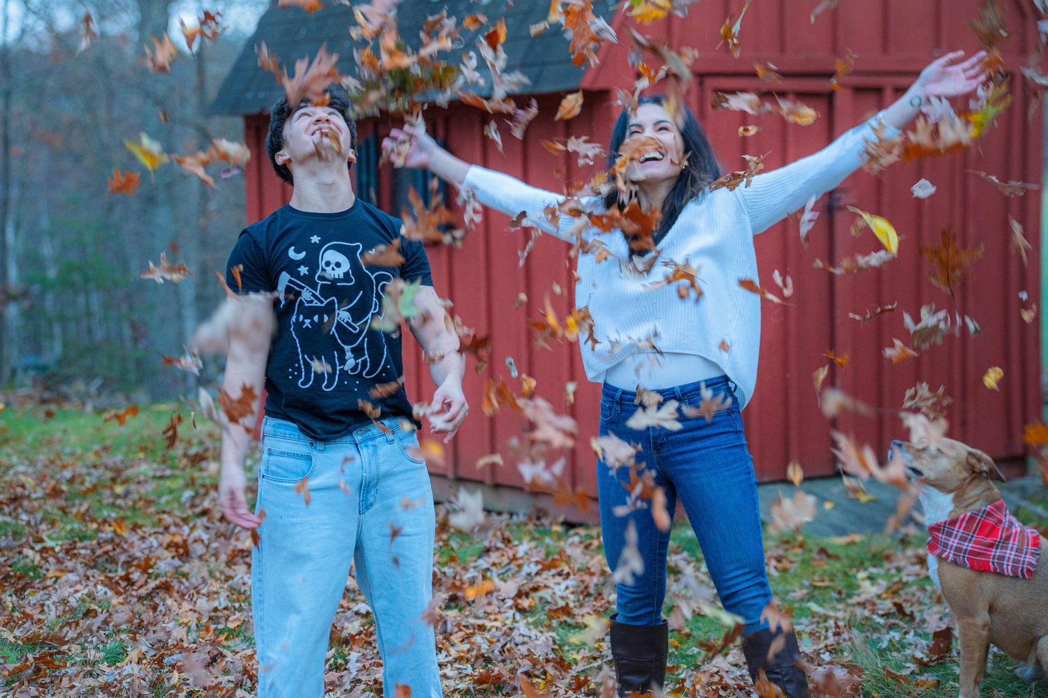 Couple joyfully tossing autumn leaves in backyard near red barn with dog, wearing casual fall outfits.