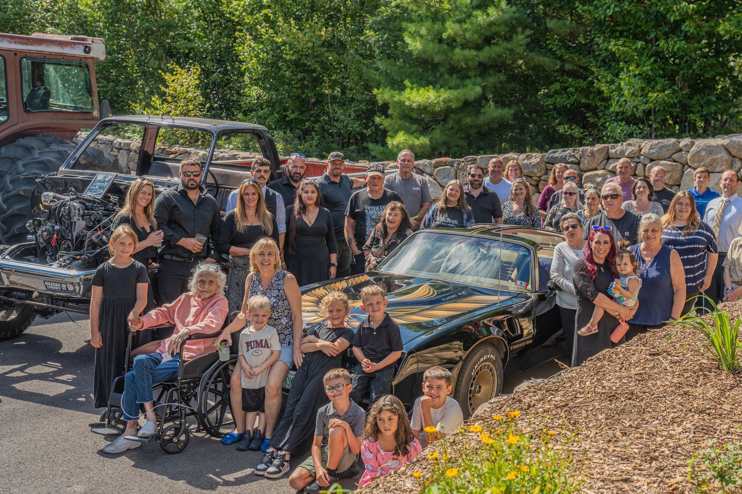 Large family group photo outdoors beside a classic black car, with trees and stone wall in background.