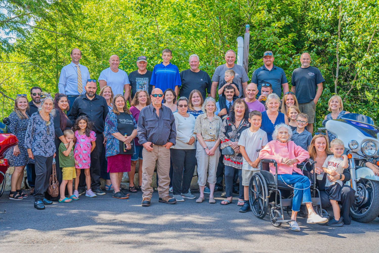 Large multigenerational family group photo outdoors, smiling together on a sunny day with lush green trees in background.