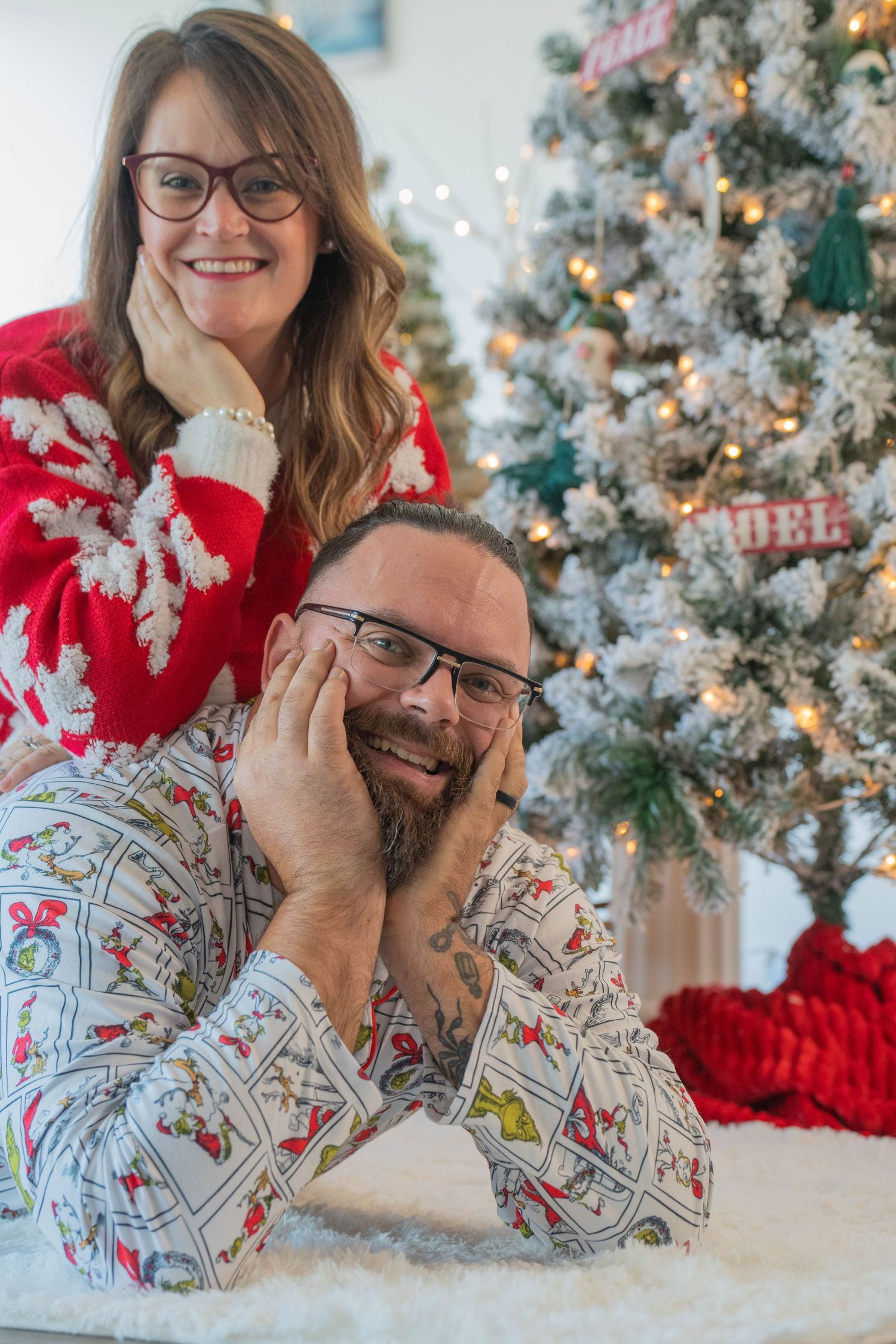 Couple in Christmas pajamas smiling by a flocked tree, woman in red snowflake sweater, man lying on white fur rug.