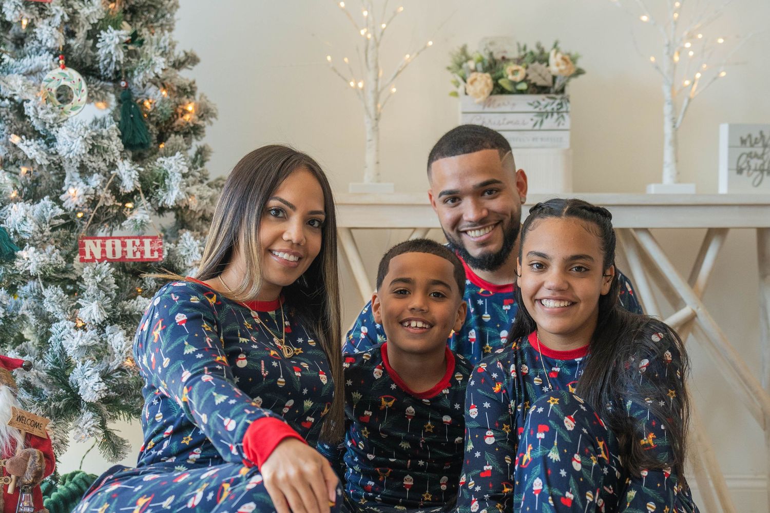 Family of four in matching blue Christmas pajamas smiling together near a decorated holiday tree.