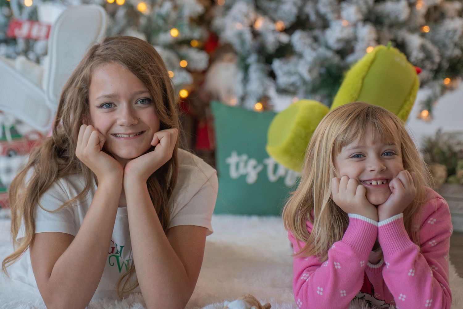 Two young girls pose cheerfully in front of a decorated Christmas tree, lying down with hands on cheeks.